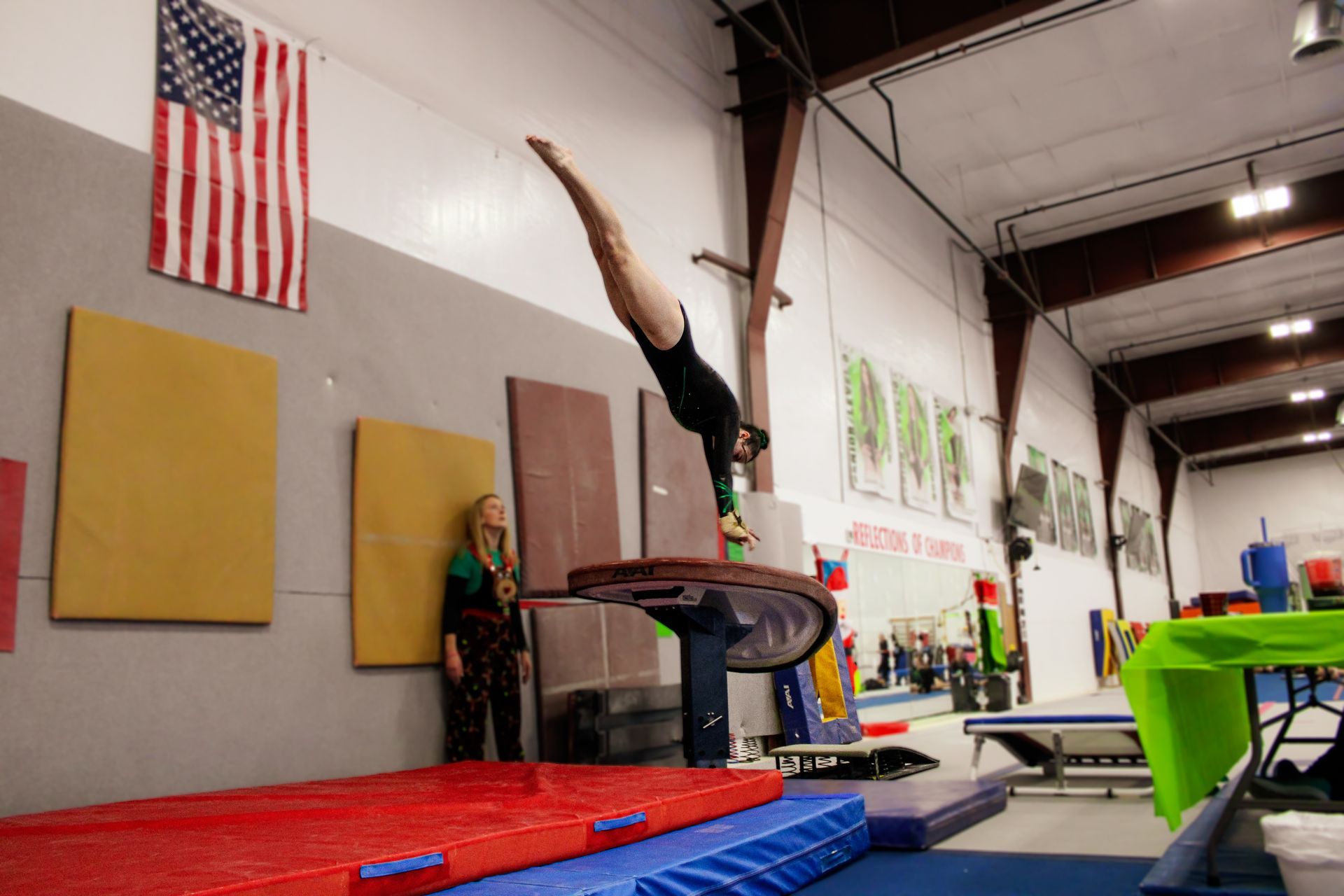 Gymnast balancing upside down on a padded beam in an indoor gym with mats and an American flag.