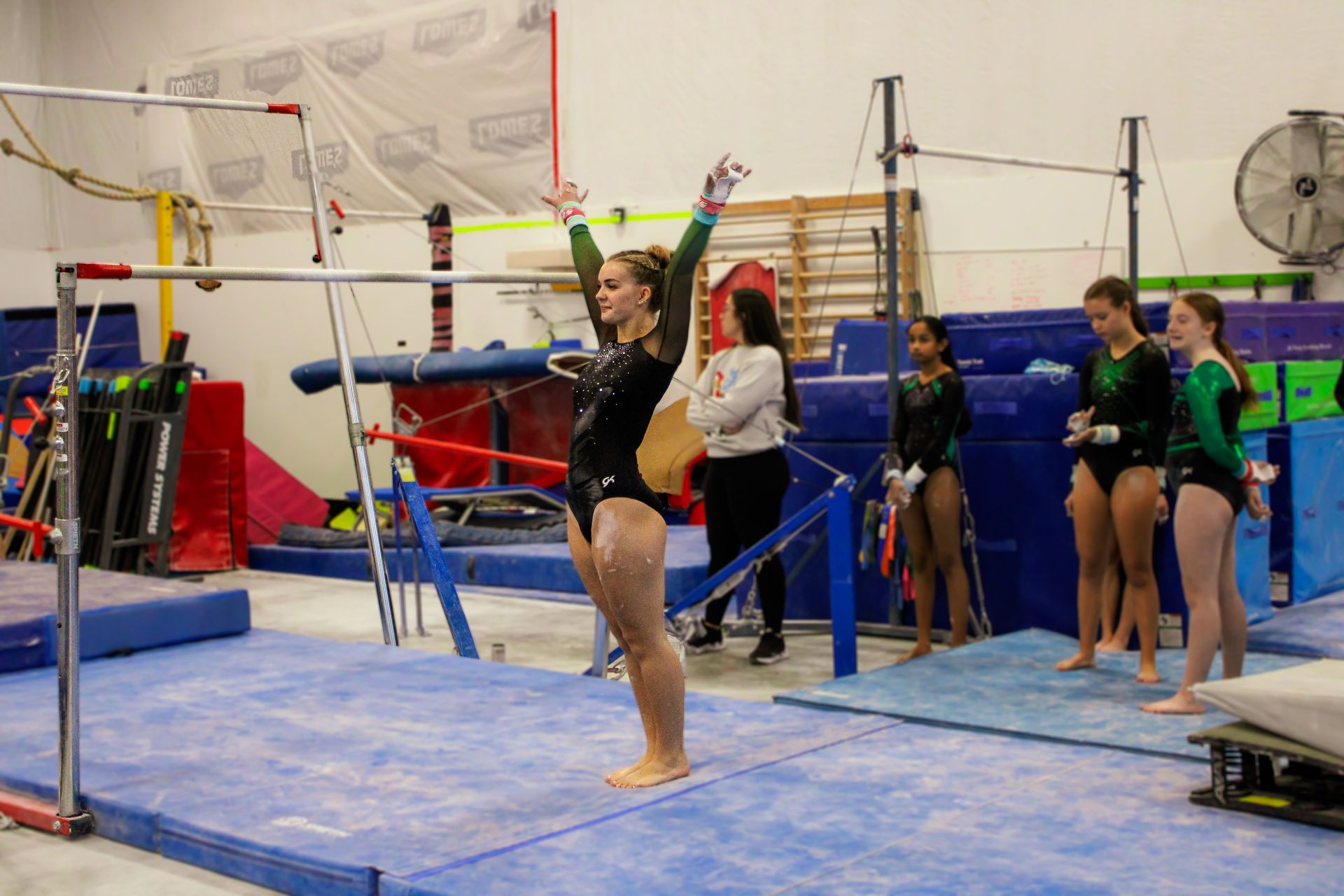 Gymnastics practice on uneven bars, with a gymnast posing on the floor and others watching in a gym.