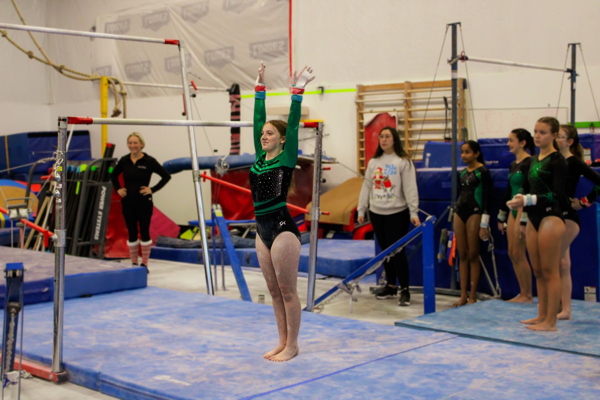 Gymnast hanging from uneven bars in a gym, with coaches and other gymnasts nearby.
