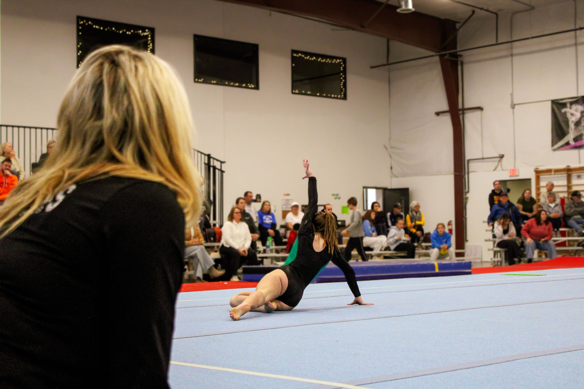 Gymnast performing a floor routine on a blue mat in a gym, with spectators watching nearby.