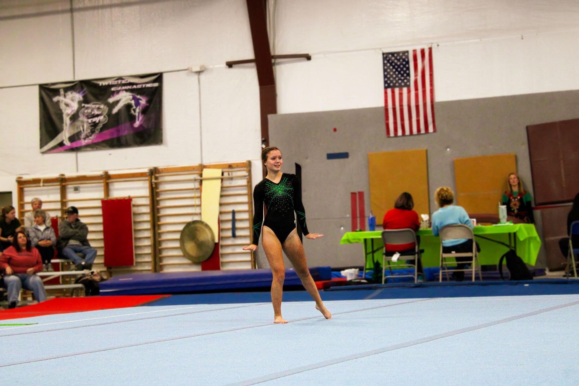 Gymnast posing on a blue mat in an indoor gym with judges and an American flag in the background