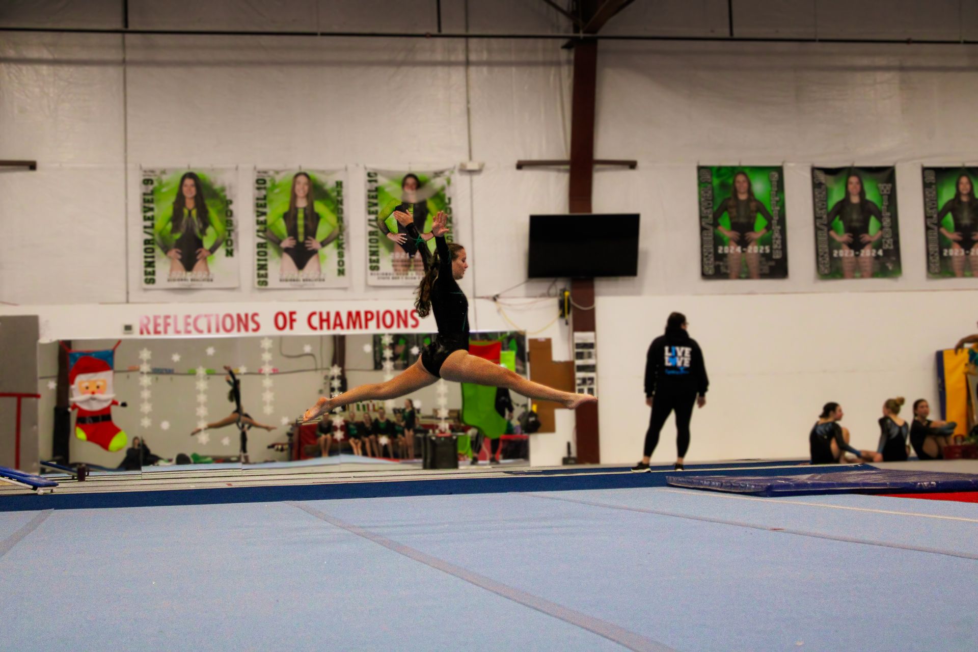 Gymnast mid-routine on balance beam in a gym, with blue mats and coaches watching nearby