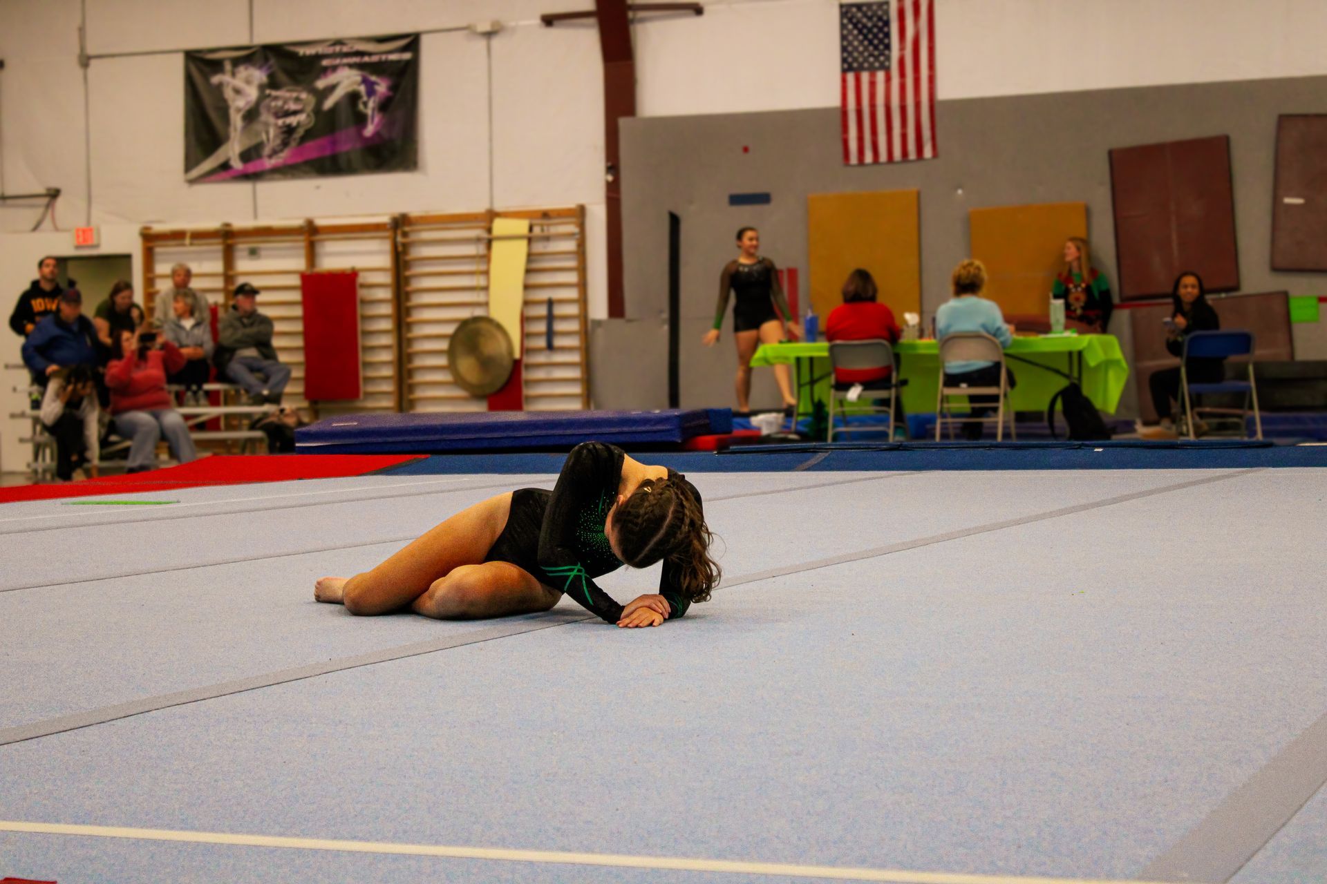 Gymnast lying on a blue mat in an indoor gymnasium, with spectators and equipment in the background