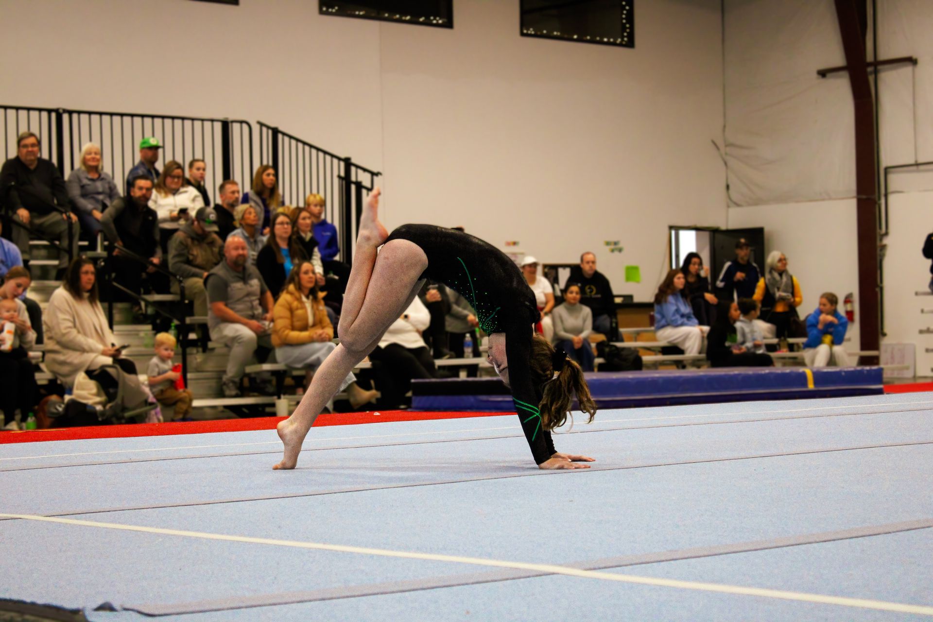 Gymnast mid-flip on a blue mat in an indoor gymnasium with spectators in the background