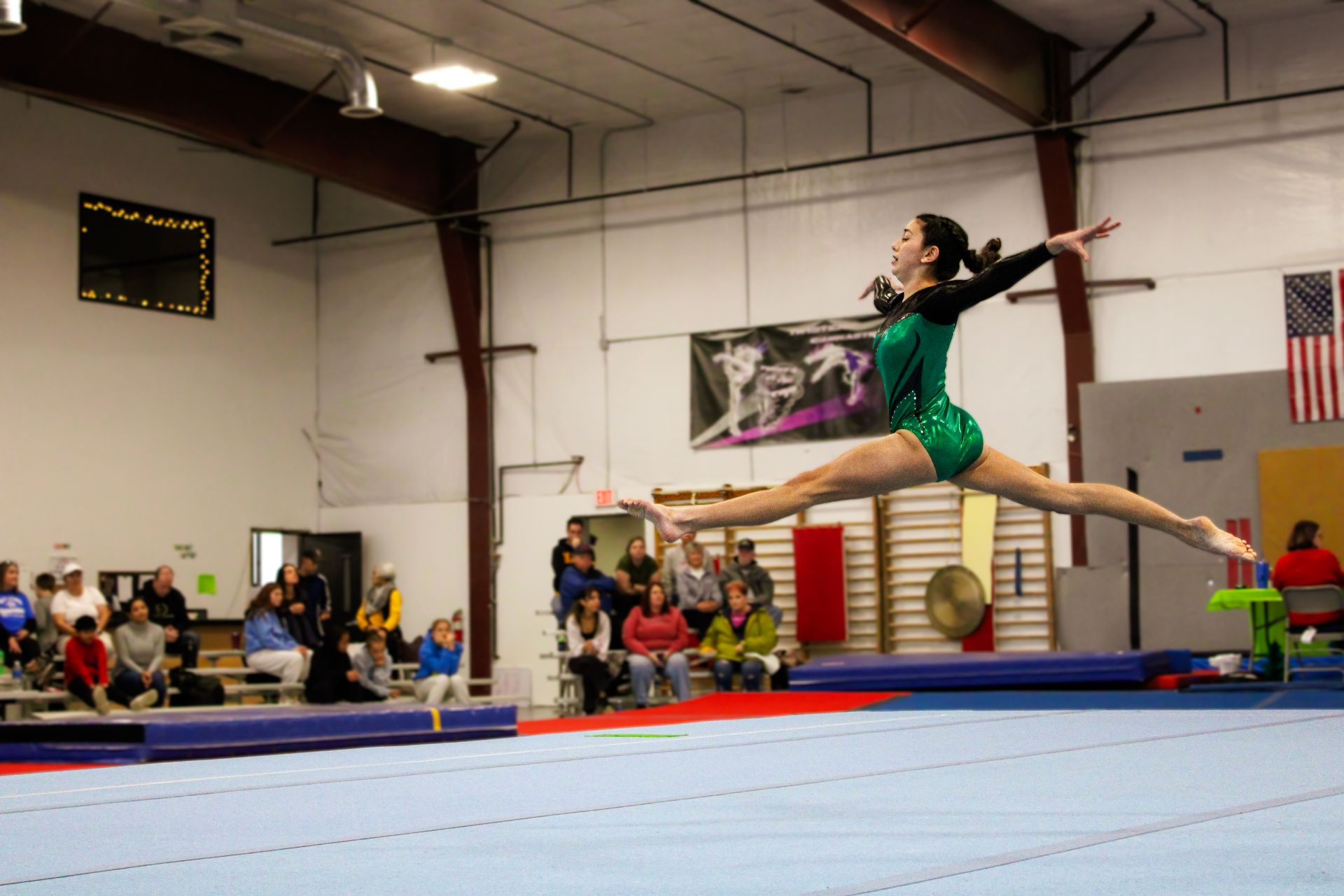 Gymnast in green leotard performing a split leap on a balance beam in an indoor gymnasium