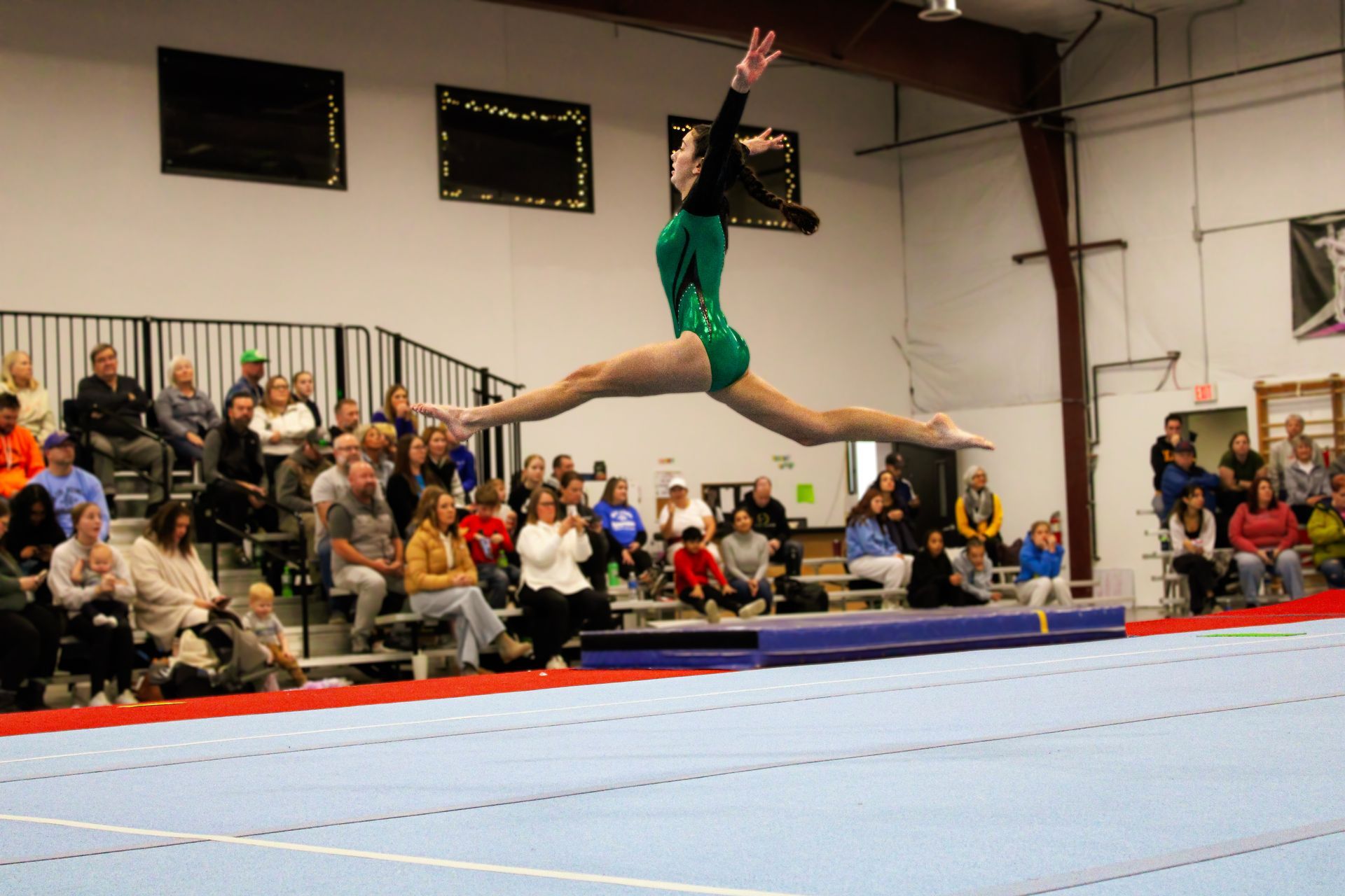 Gymnast in green leotard leaps across a blue floor mat in a gymnasium with spectators watching.