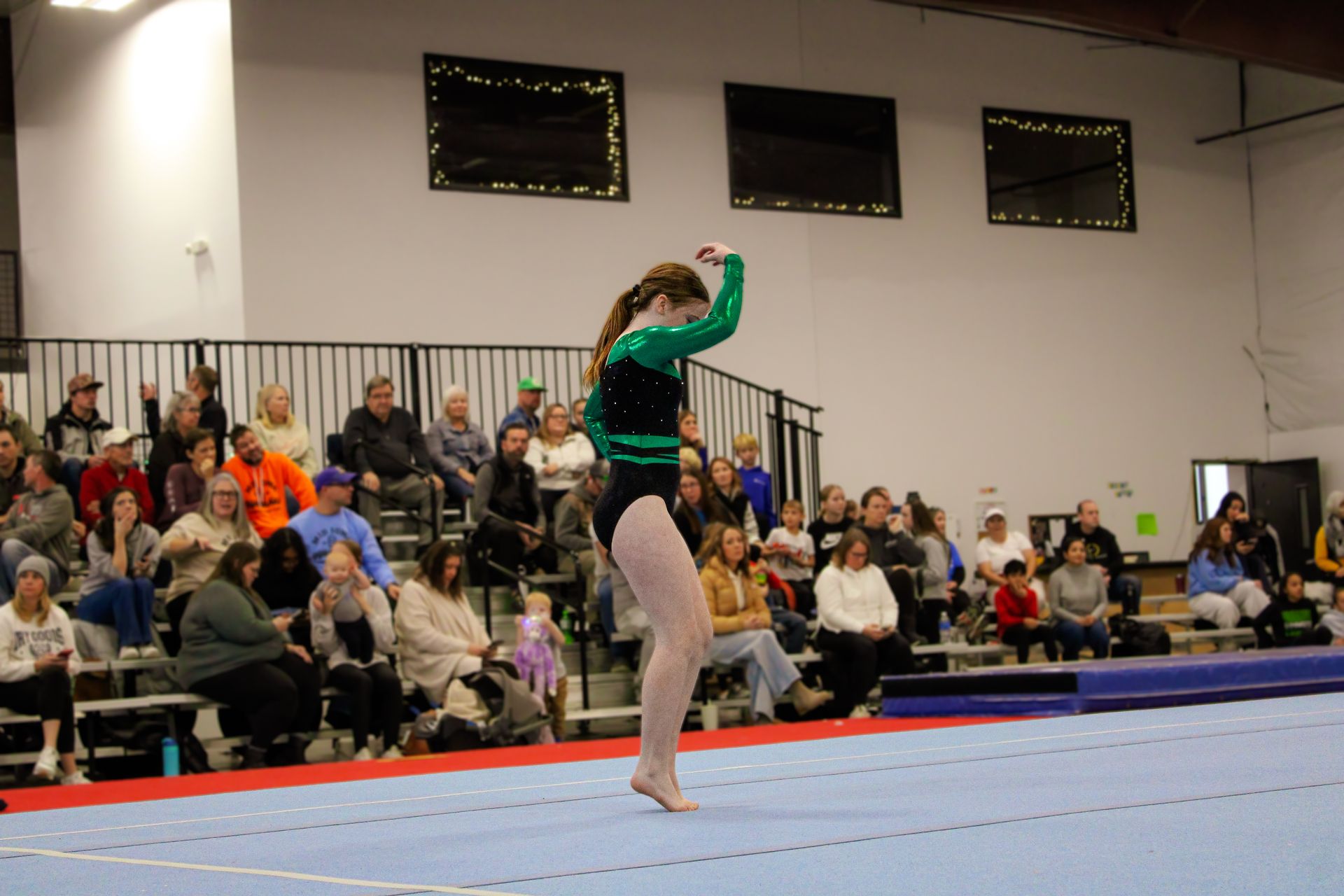 Gymnast in a green leotard performing on a mat before a seated audience indoors