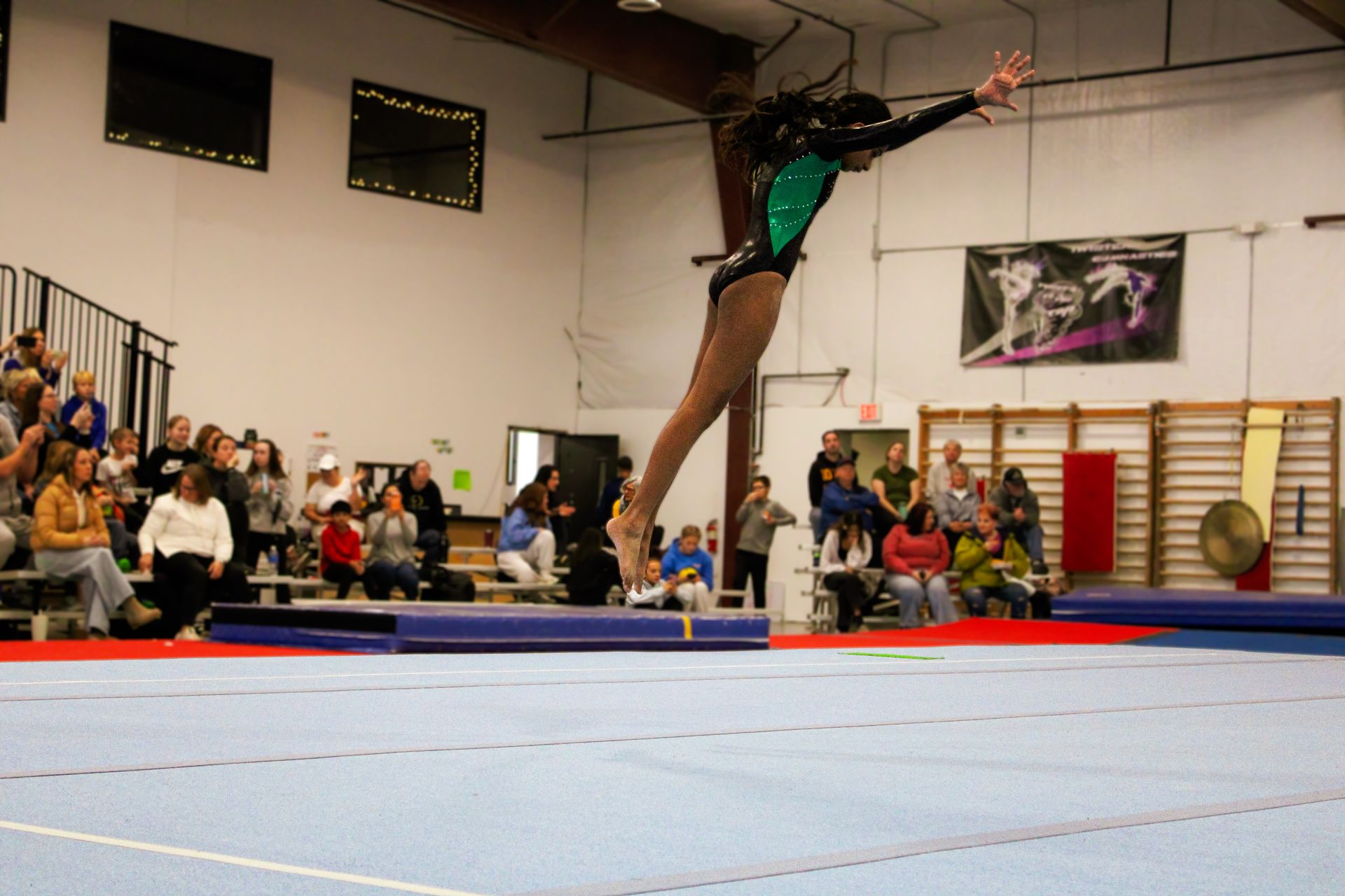 Gymnast performing a leap on a blue mat in an indoor gym with spectators watching