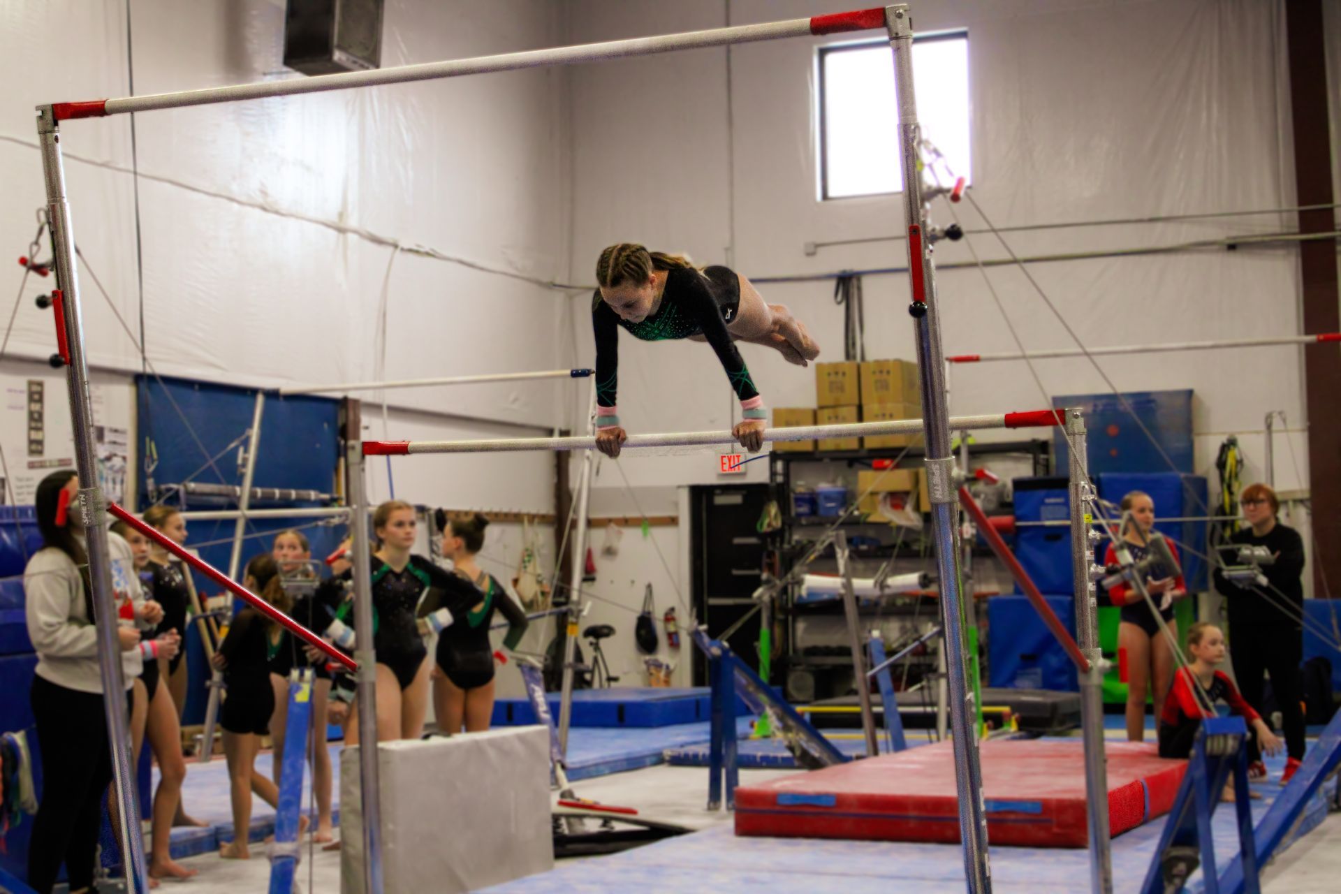 Gymnast mid-air over uneven bars in a gym, with spectators and safety mats below