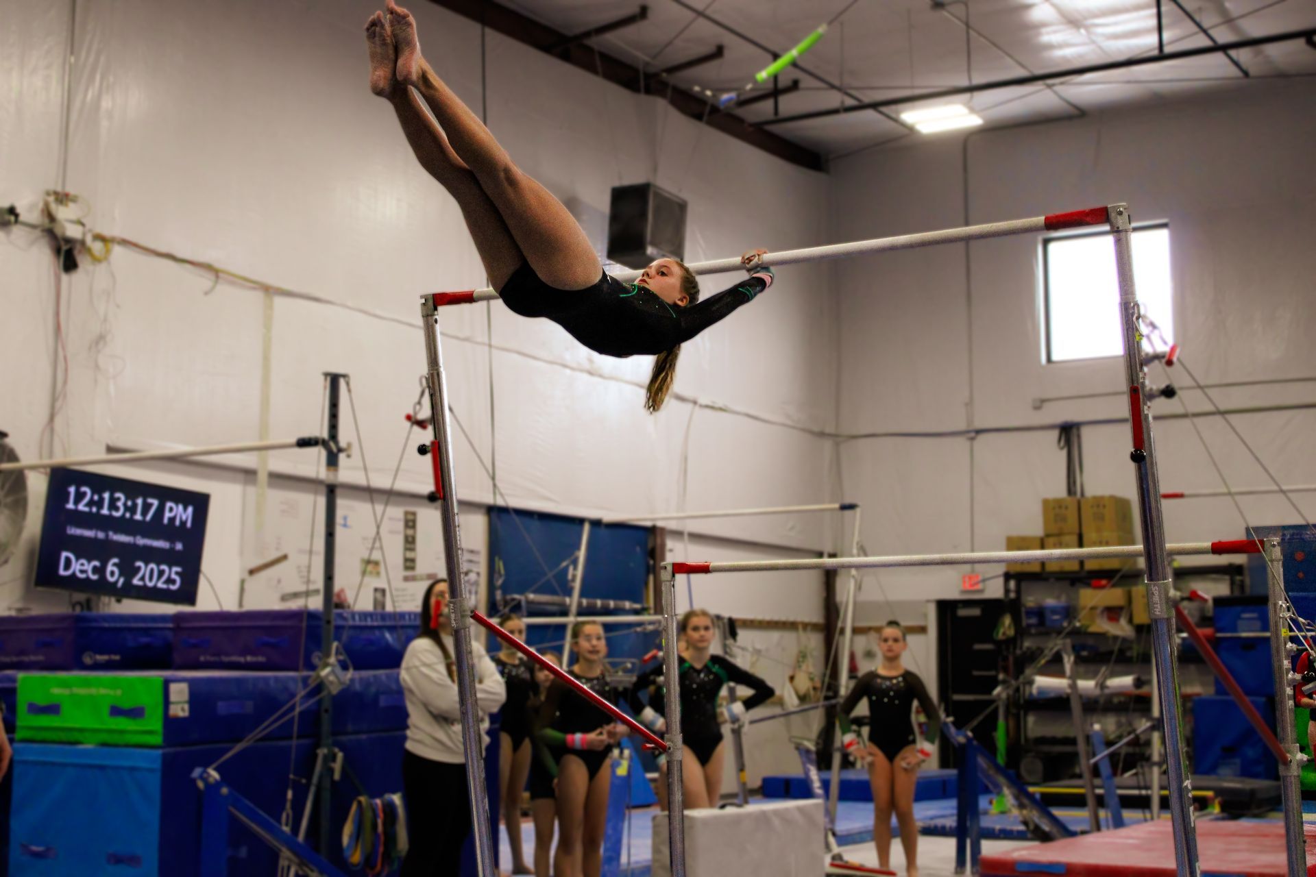 Gymnast performing a horizontal bar swing in an indoor gymnastics gym, with coaches and athletes watching nearby.