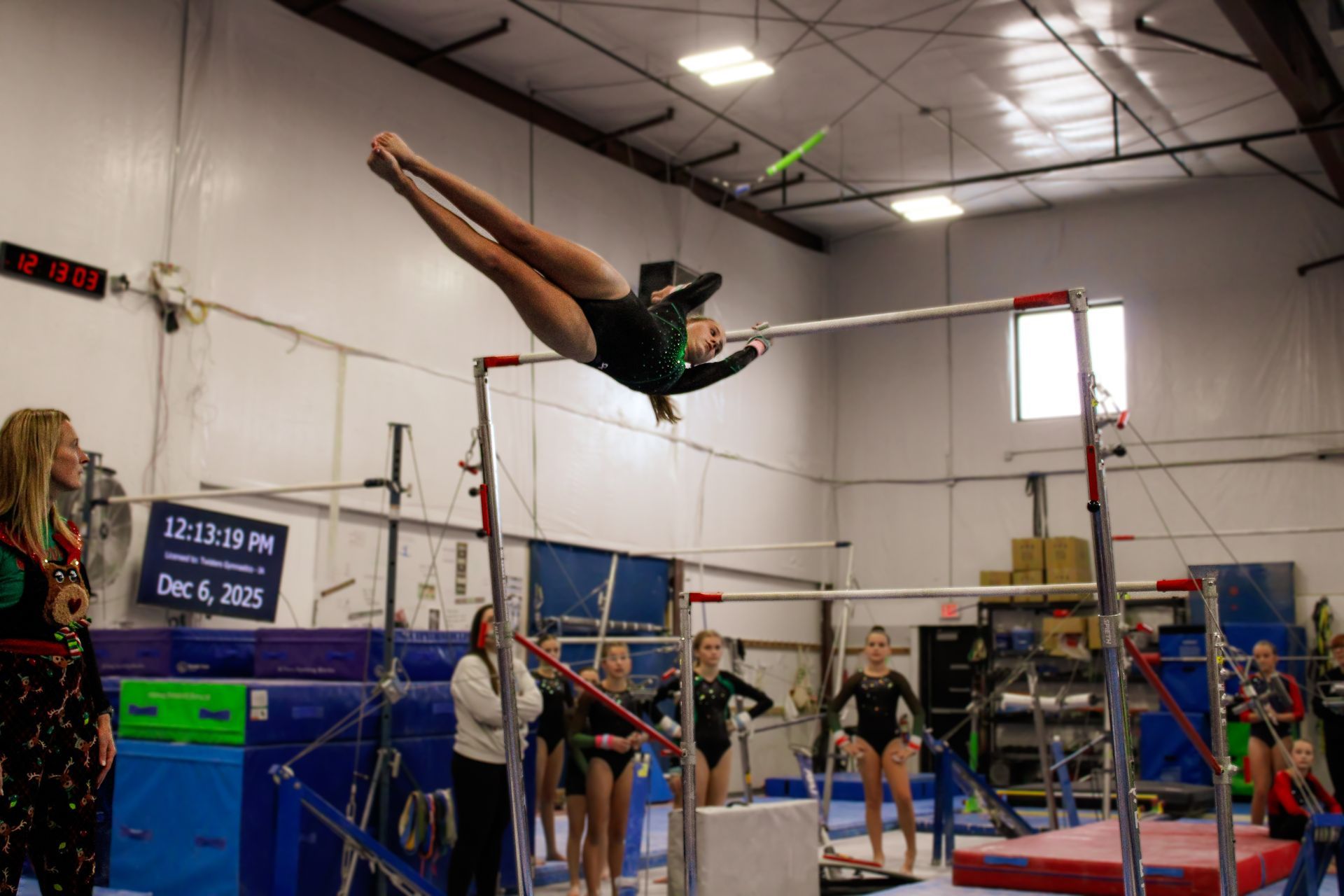 Gymnast performing a high bar release move in a gym, with coaches and other athletes watching nearby.
