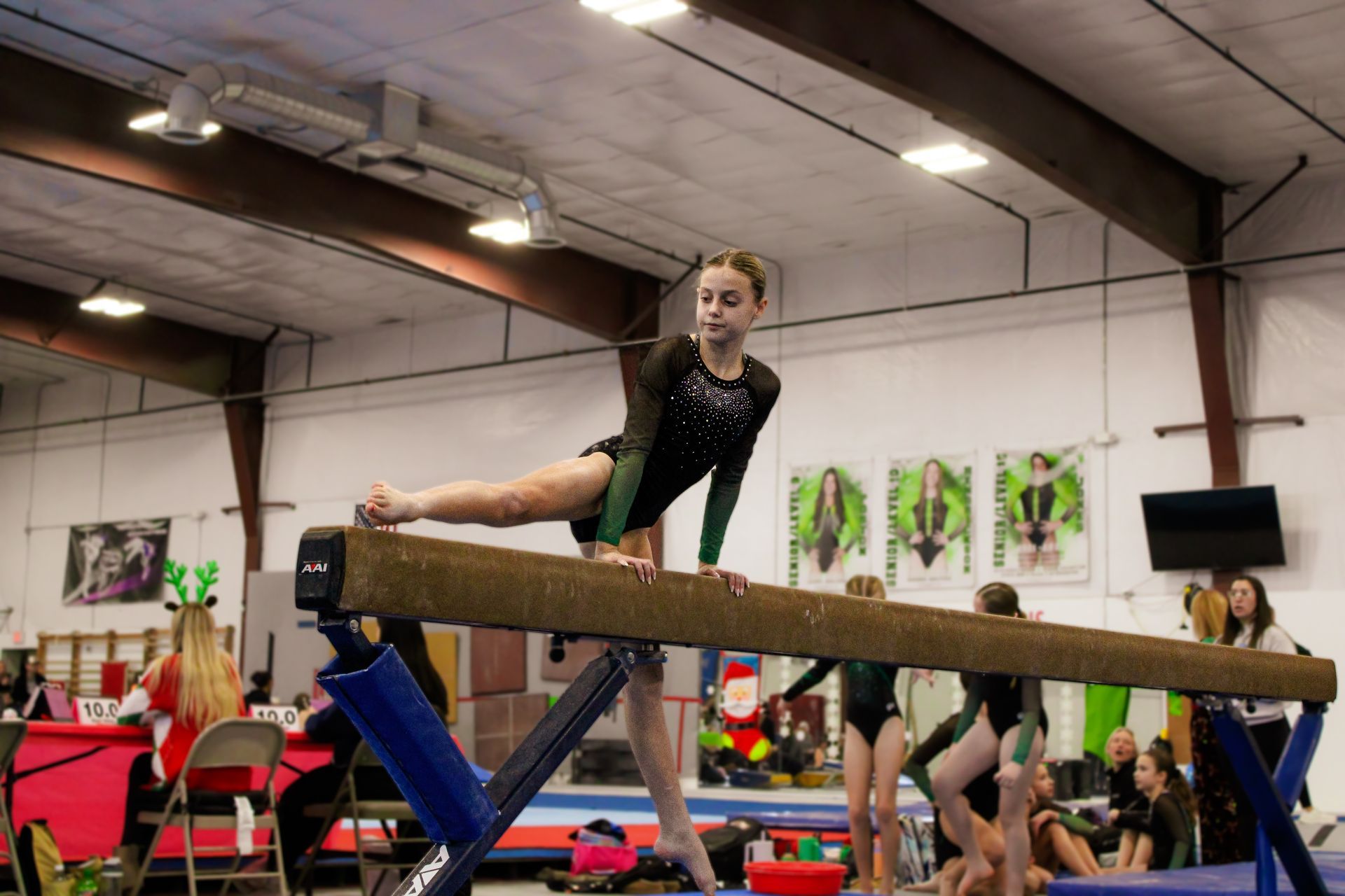 Gymnast balancing on a beam in an indoor training facility, with coaches and equipment in the background