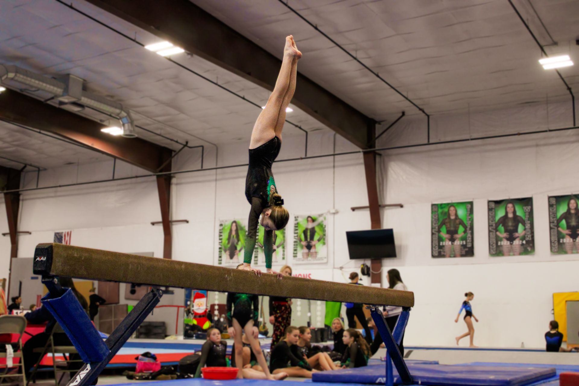 Gymnast upside down on a balance beam in a gymnasium, with spectators and posters in the background.