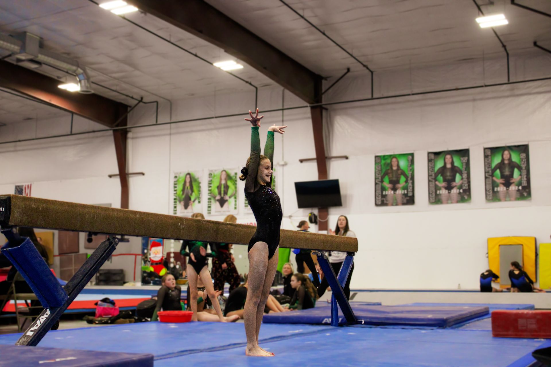 Gymnast balancing on one foot with arms raised beside a balance beam in a gymnasium