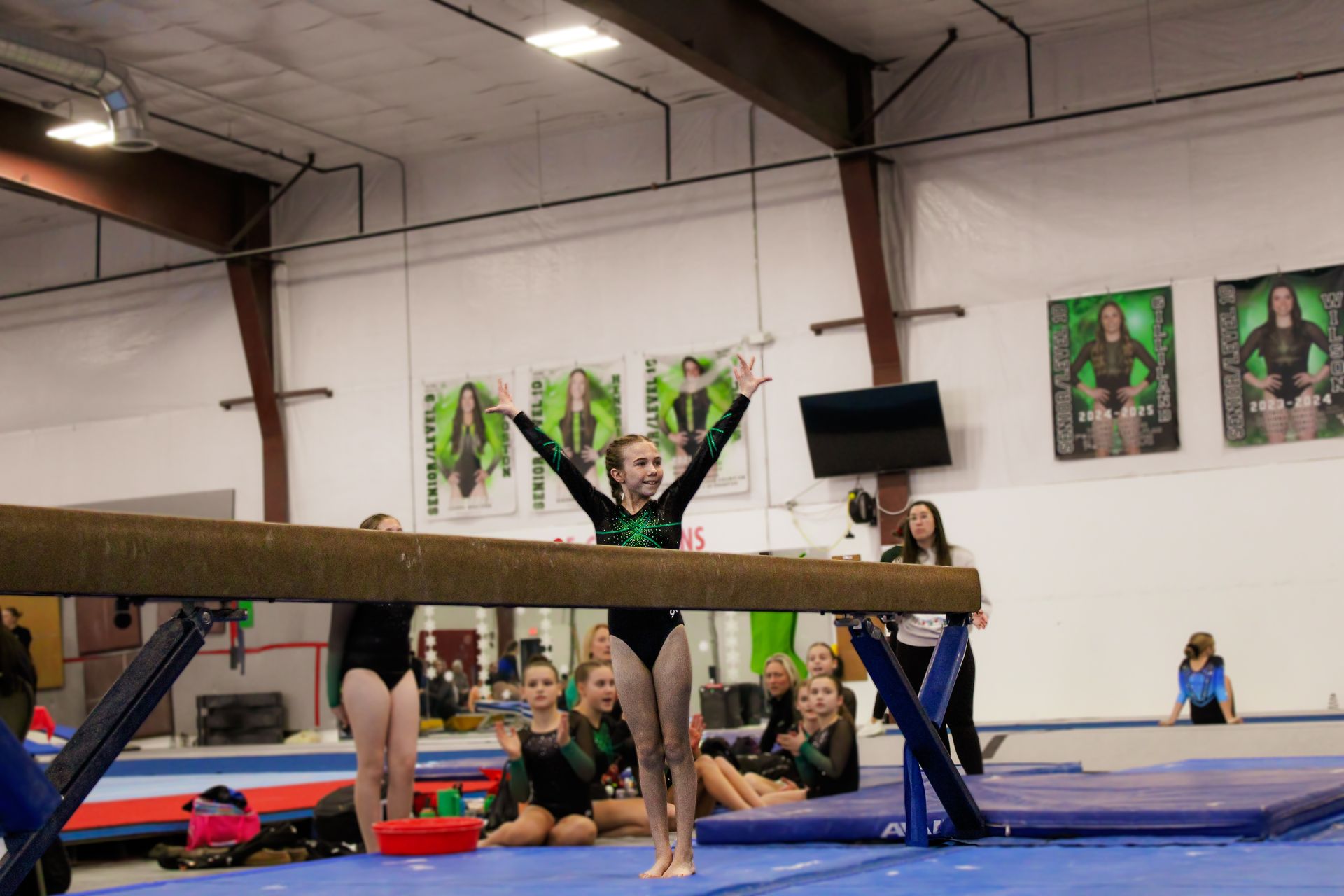 Gymnast balancing on a beam in a gym, with teammates watching in the background.