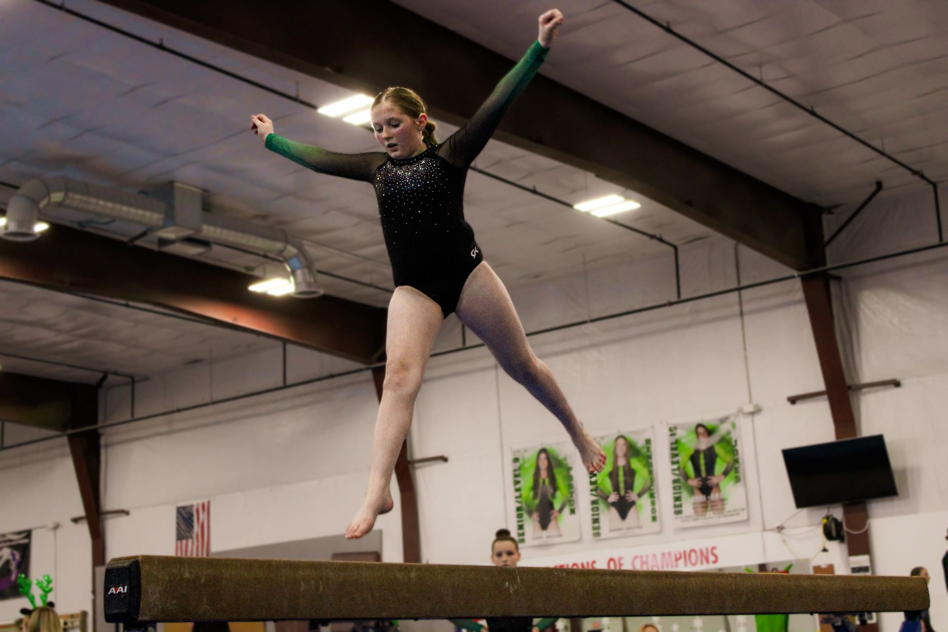 Gymnast balancing on a beam indoors with arms raised in a black leotard.