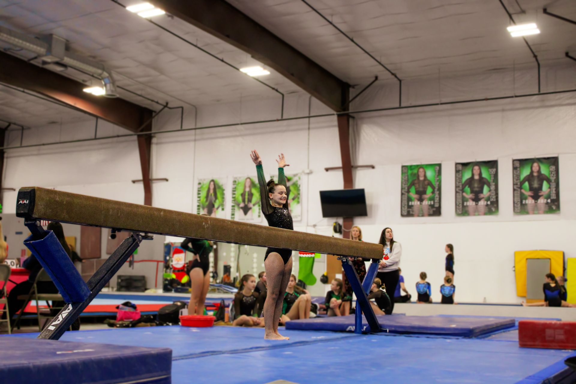 Gymnast balancing on a beam in an indoor gym, with other athletes and mats in the background.