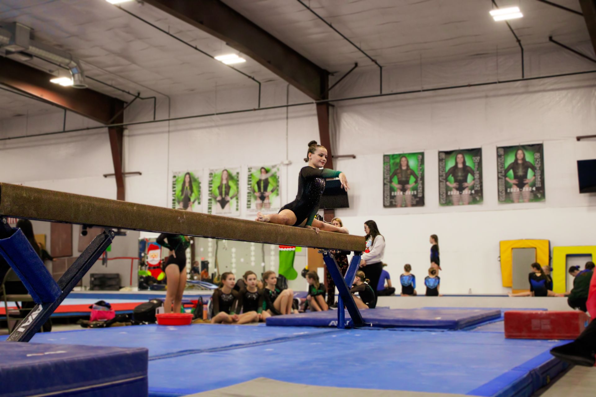 Gymnast balancing on a beam in a gymnasium, with spectators and posters in the background.