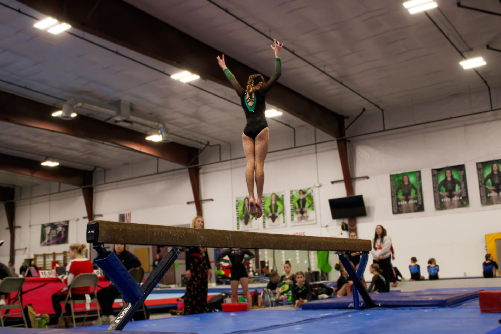Gymnast balancing on a beam in an indoor gym, arms raised, with a spotting mat below