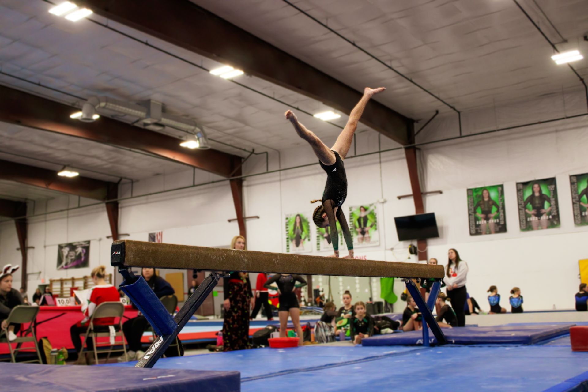 Gymnast performing a handstand on a balance beam in a gymnasium, with mats and spectators nearby