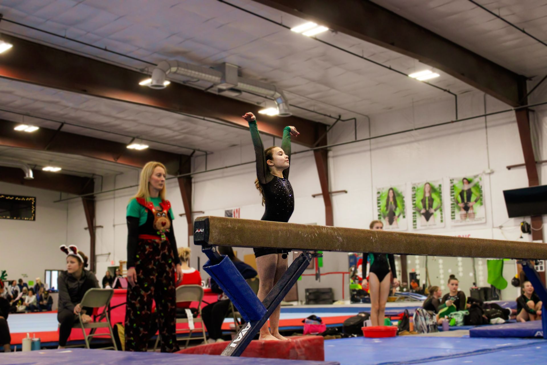 Gymnast balancing on a beam in an indoor gym, with a coach nearby and spectators in the background