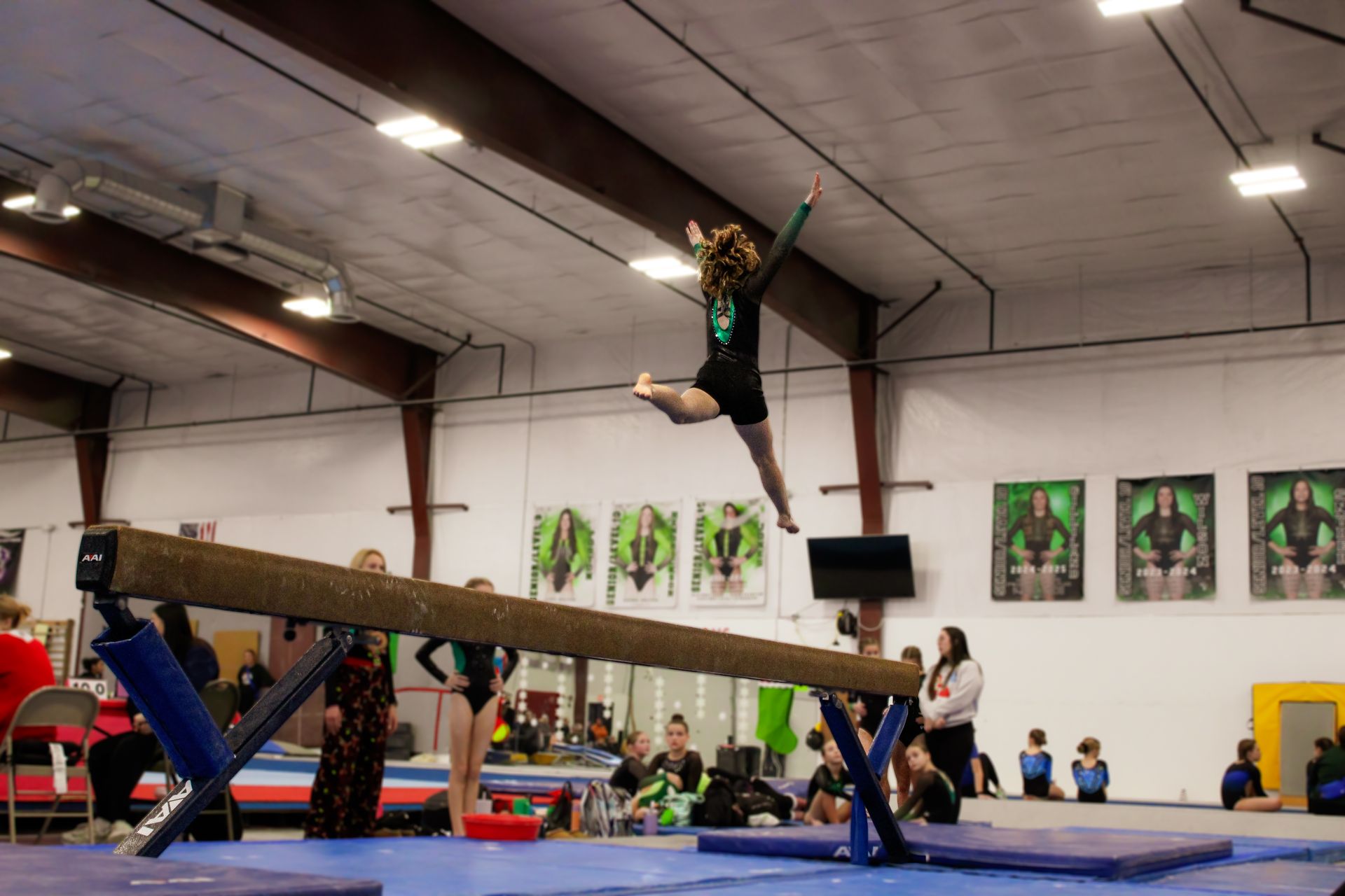 Gymnast mid-air over balance beam in an indoor gymnasium, arms and legs extended during a leap.
