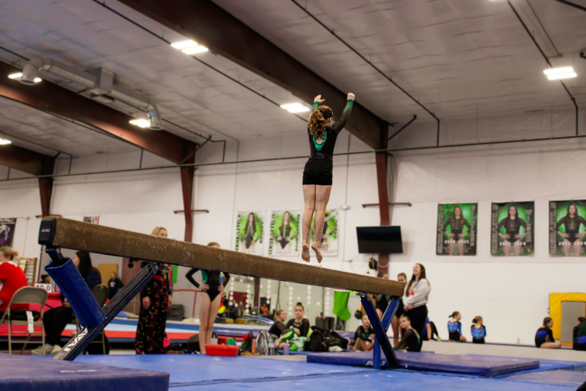 Gymnast balancing on a beam in a gymnasium during practice, with mats and spectators nearby.