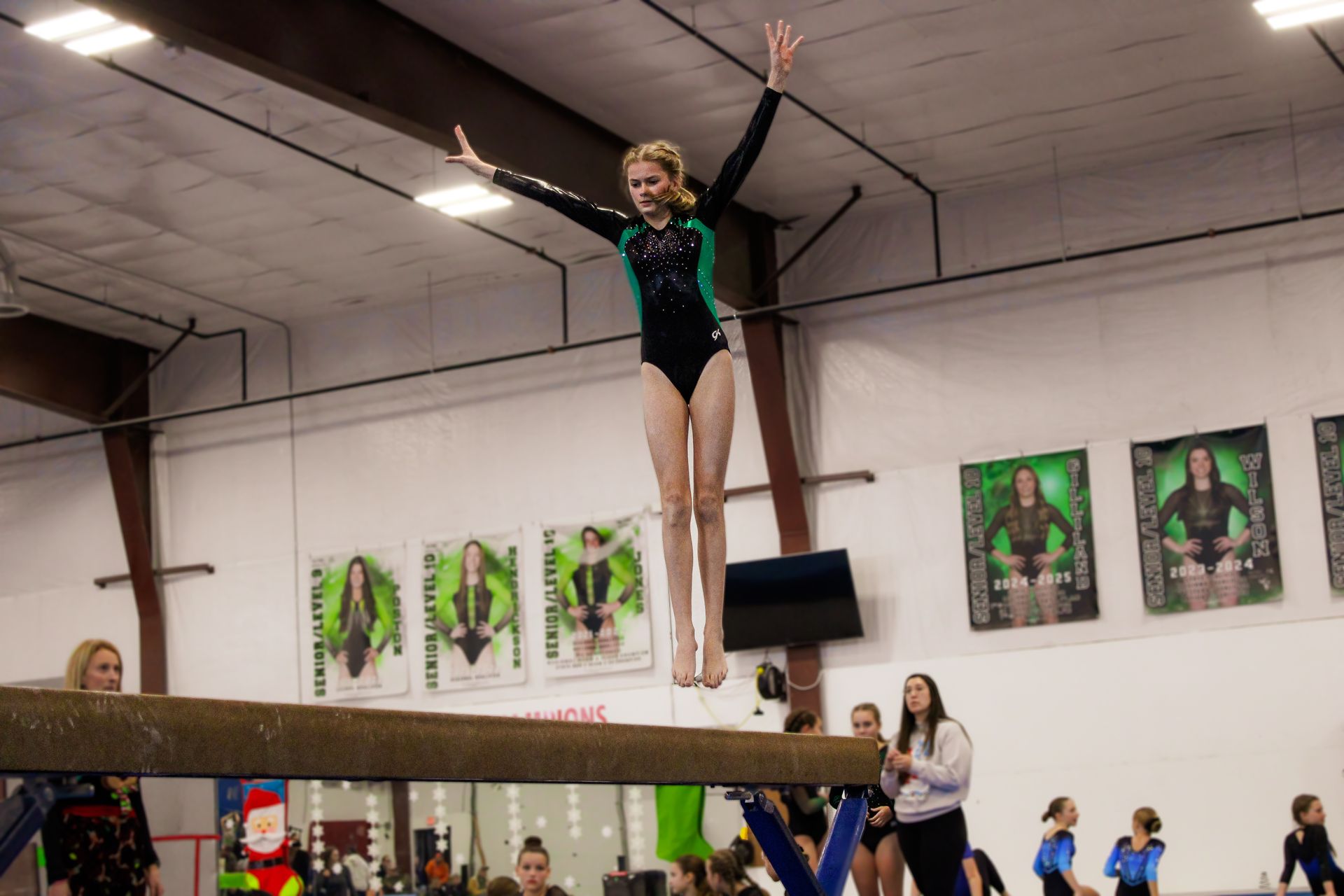 Gymnast balancing on a beam in a gym, arms raised, with spectators in the background