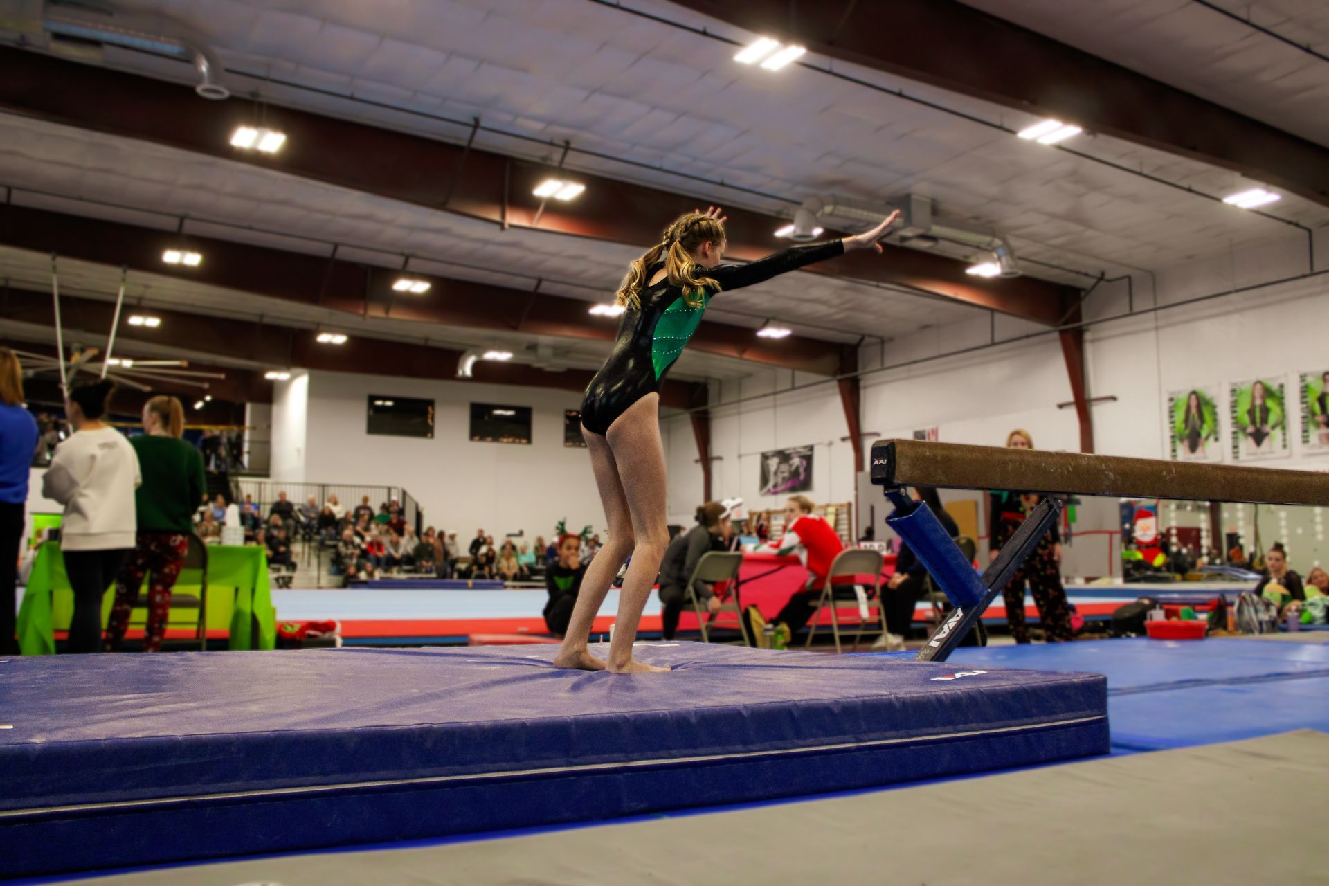 Gymnast balancing on a blue mat in an indoor gym, with judges and spectators in the background