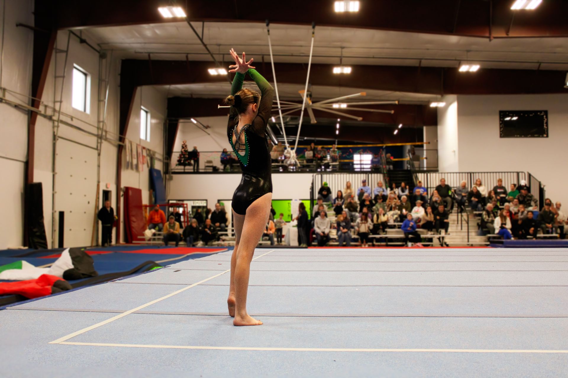 Gymnast in a black leotard balancing on a beam in an indoor competition gym, spectators in the background