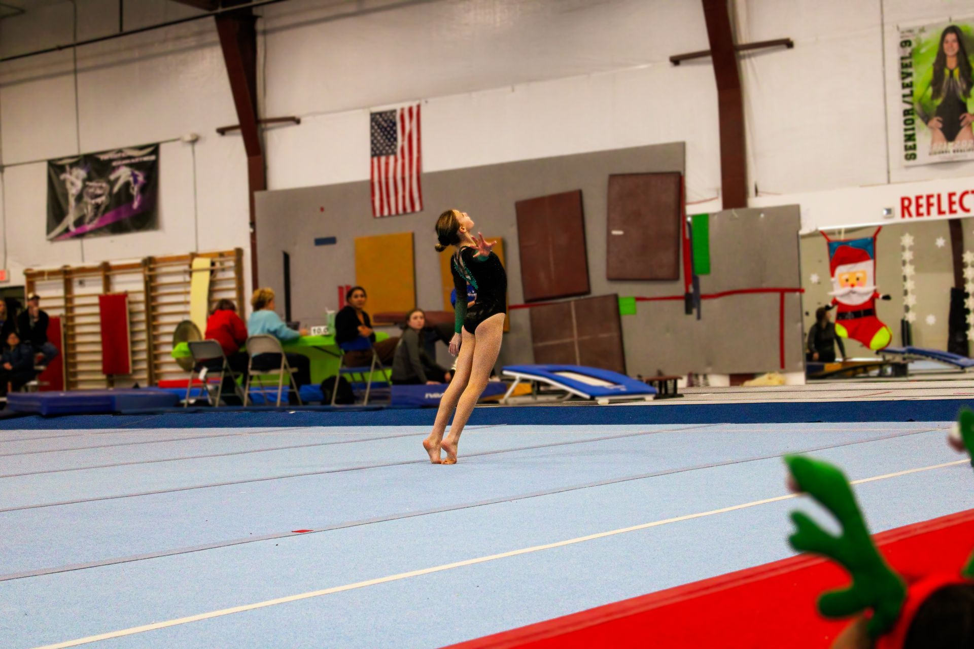 Gymnast balancing on a blue mat in an indoor gym, with judges and flags in the background