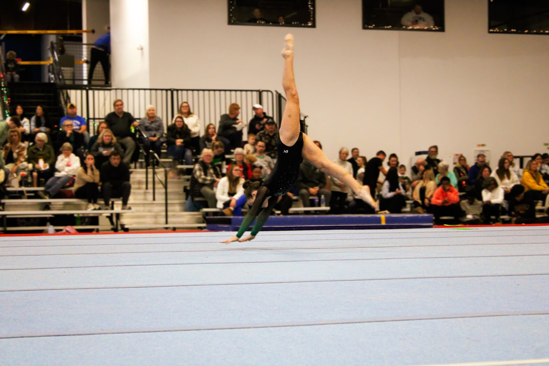 Gymnast performing a one-handed handstand on a blue mat with spectators in bleachers behind her