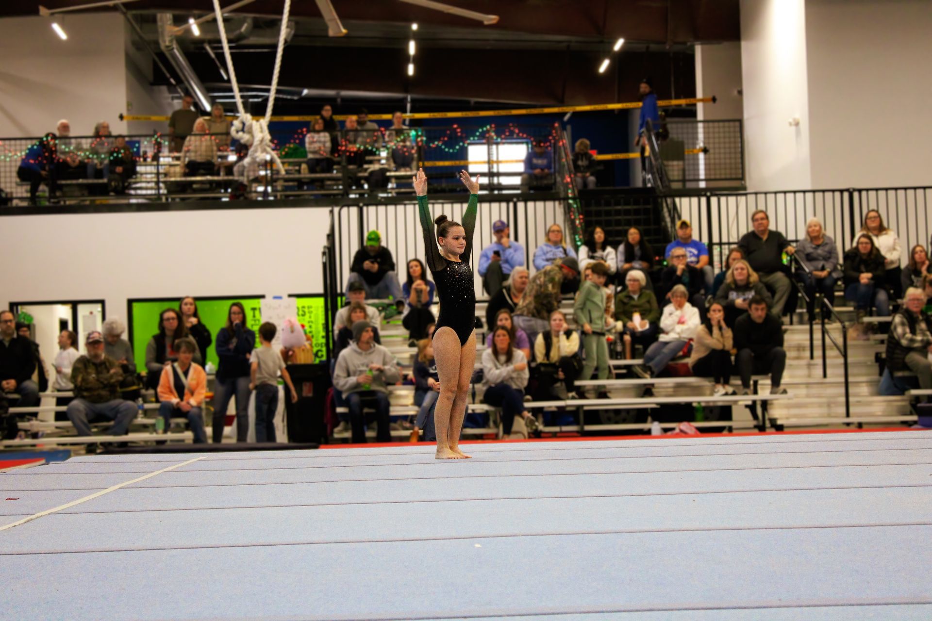 Gymnast standing on a blue mat in a gym, with spectators seated in the background.