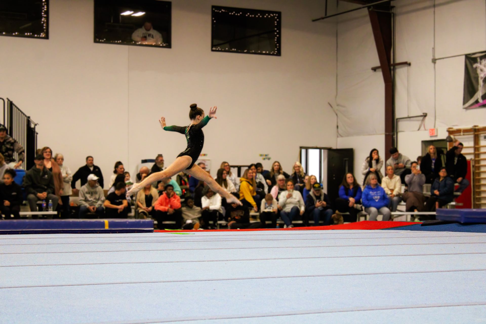Gymnast mid-air on a blue mat in an indoor arena, with spectators watching from the sidelines.