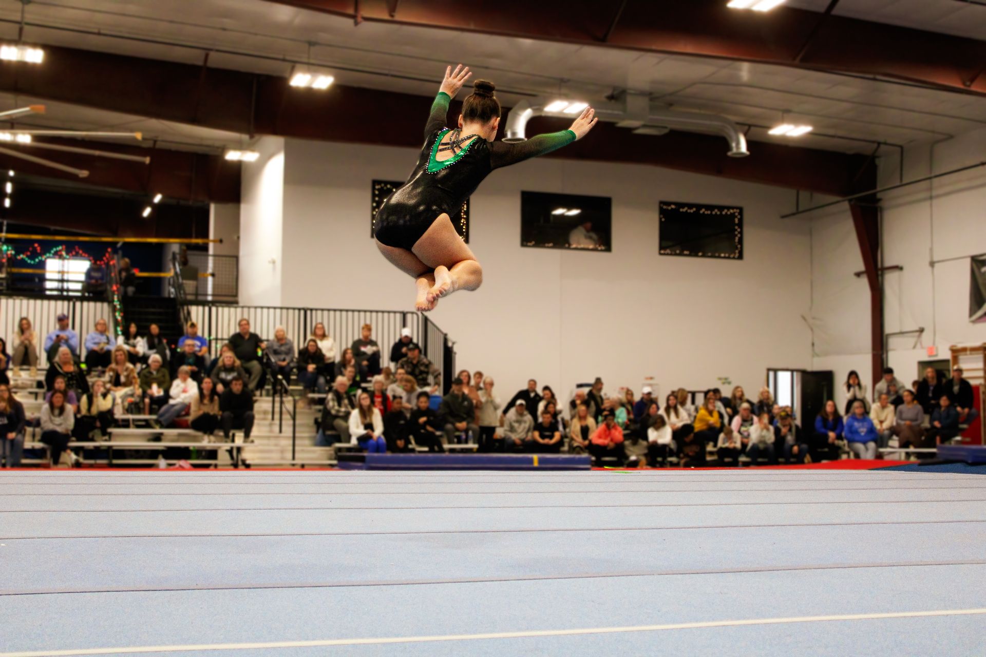 Gymnast mid-air during a vault in an indoor arena, with spectators watching in the background