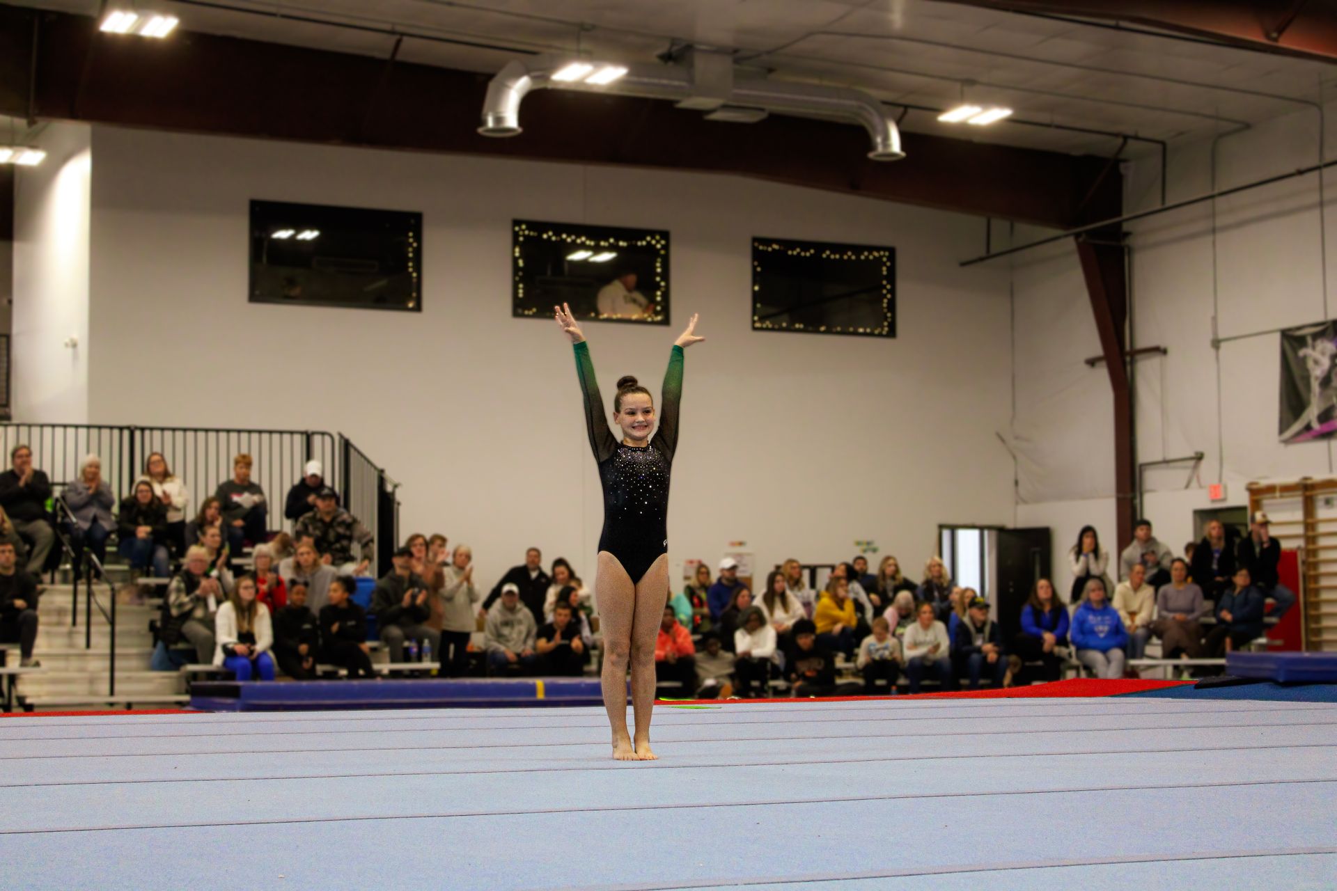 Gymnast performing a floor routine in a gymnasium, arms raised, with spectators seated in the background
