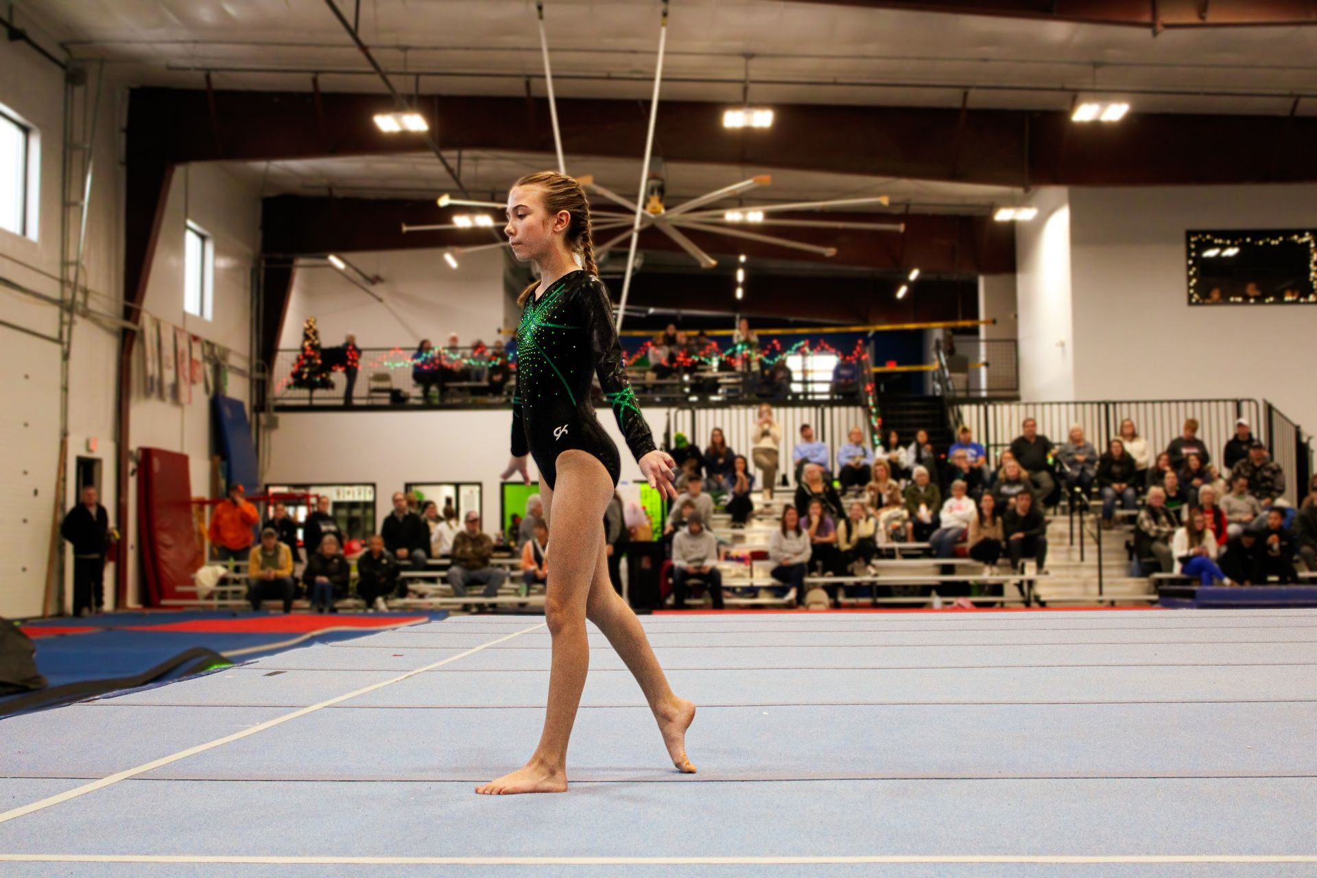 Gymnast in a black leotard walking on a floor exercise mat in an indoor gymnasium.
