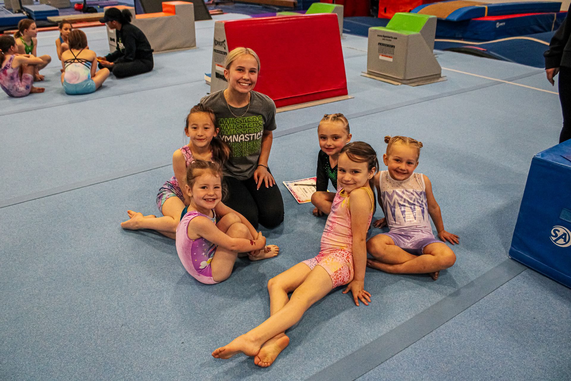 Children sitting on a blue gym mat, smiling in a play area with colorful padded blocks.
