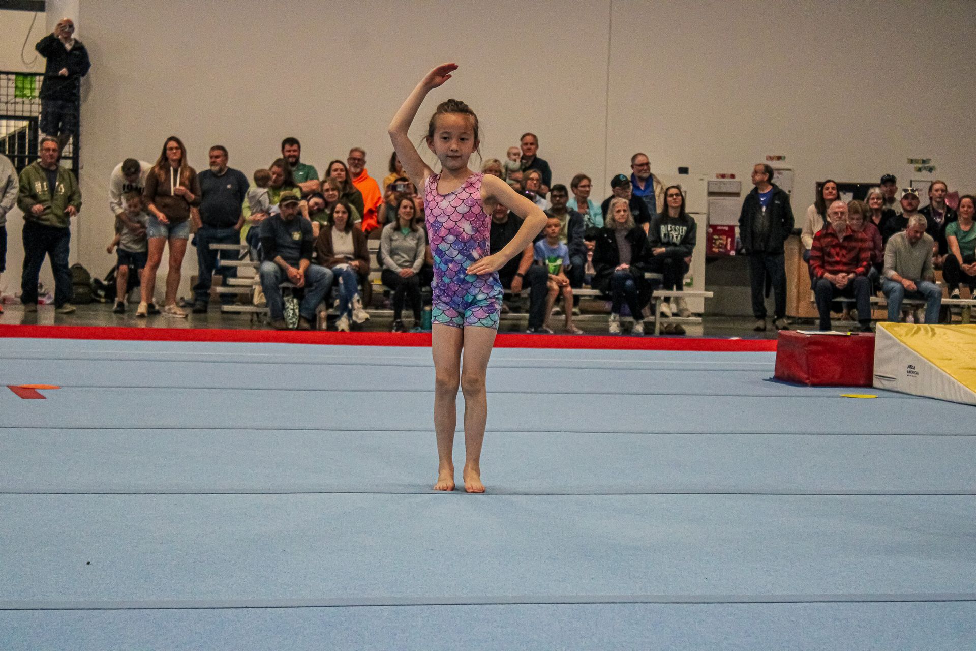 Gymnast standing on a mat with one arm raised, with spectators seated in the background.