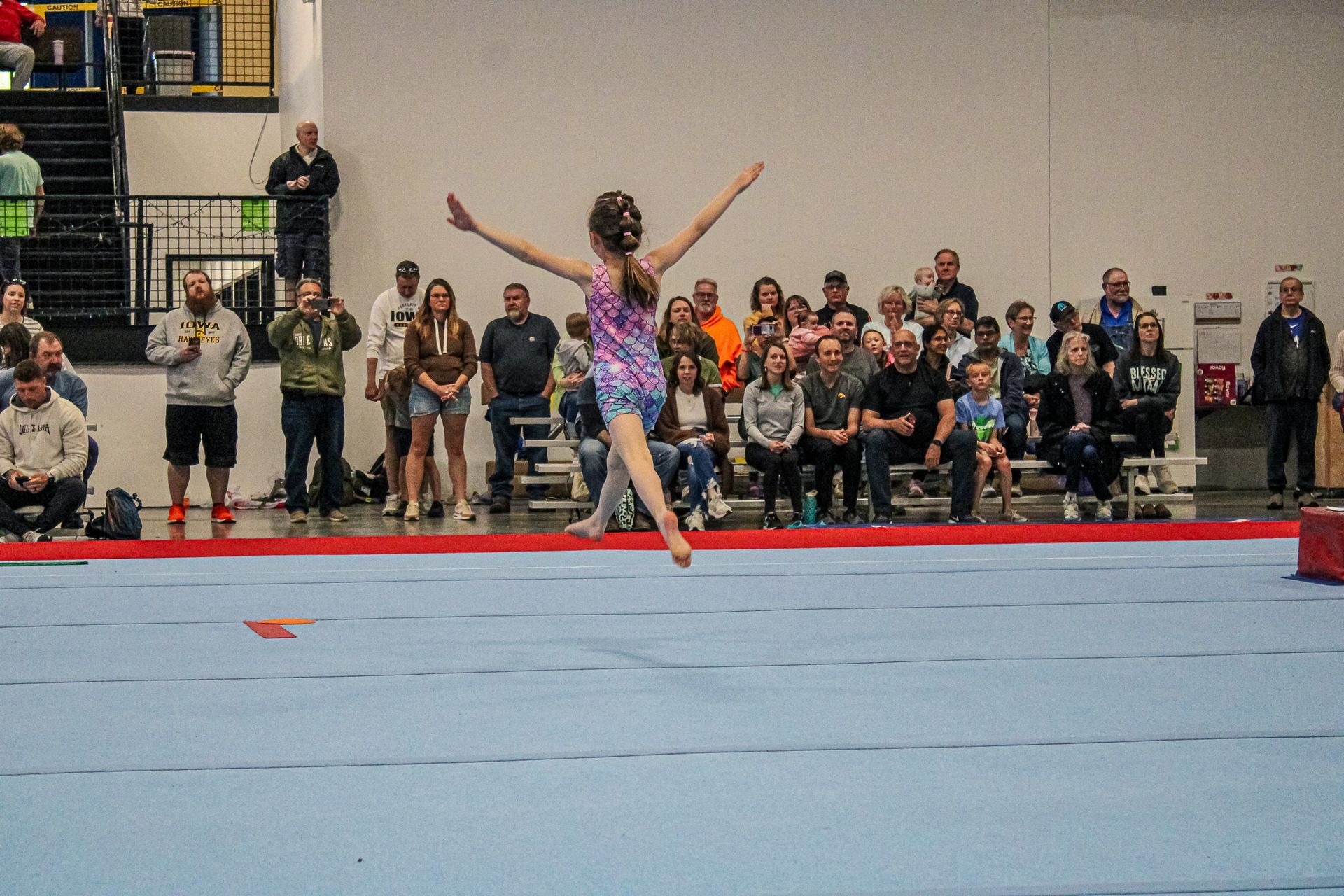 Gymnast mid-leap on a blue floor mat in a gym, with spectators watching in the background.