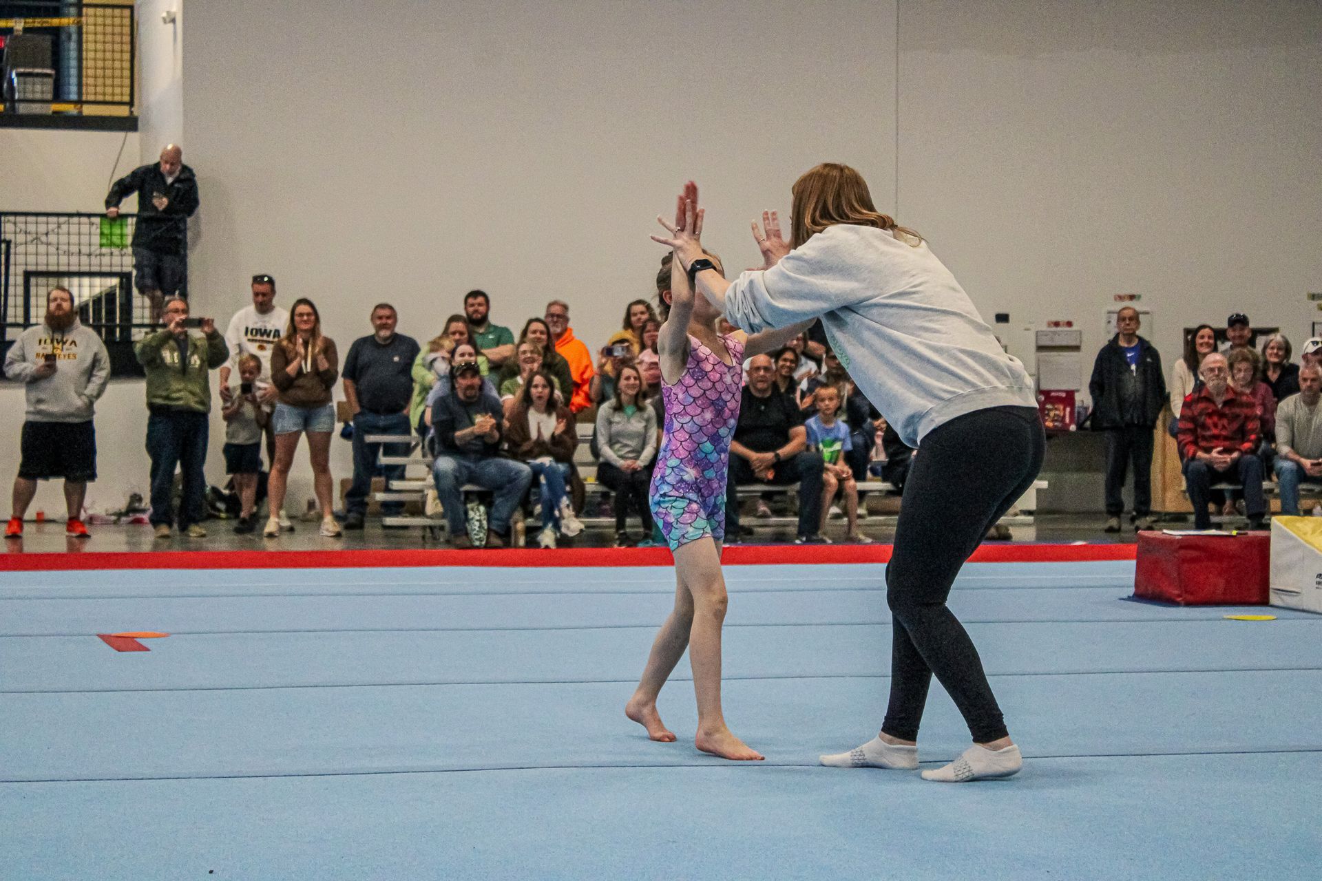 Gymnastics coach and young gymnast practicing a balance pose on a blue mat before spectators