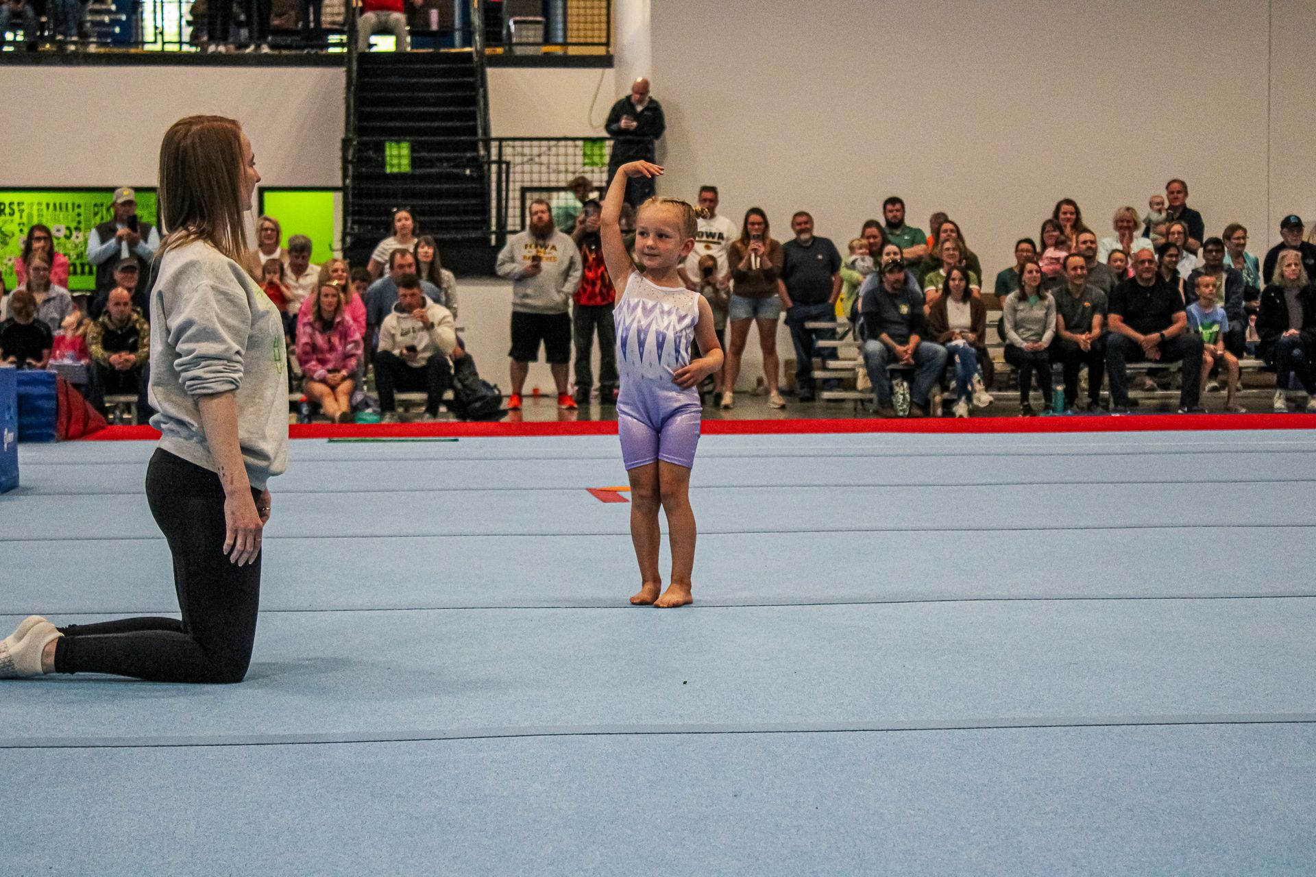 Gymnast performing on a blue mat while a judge kneels nearby, with spectators watching in the background