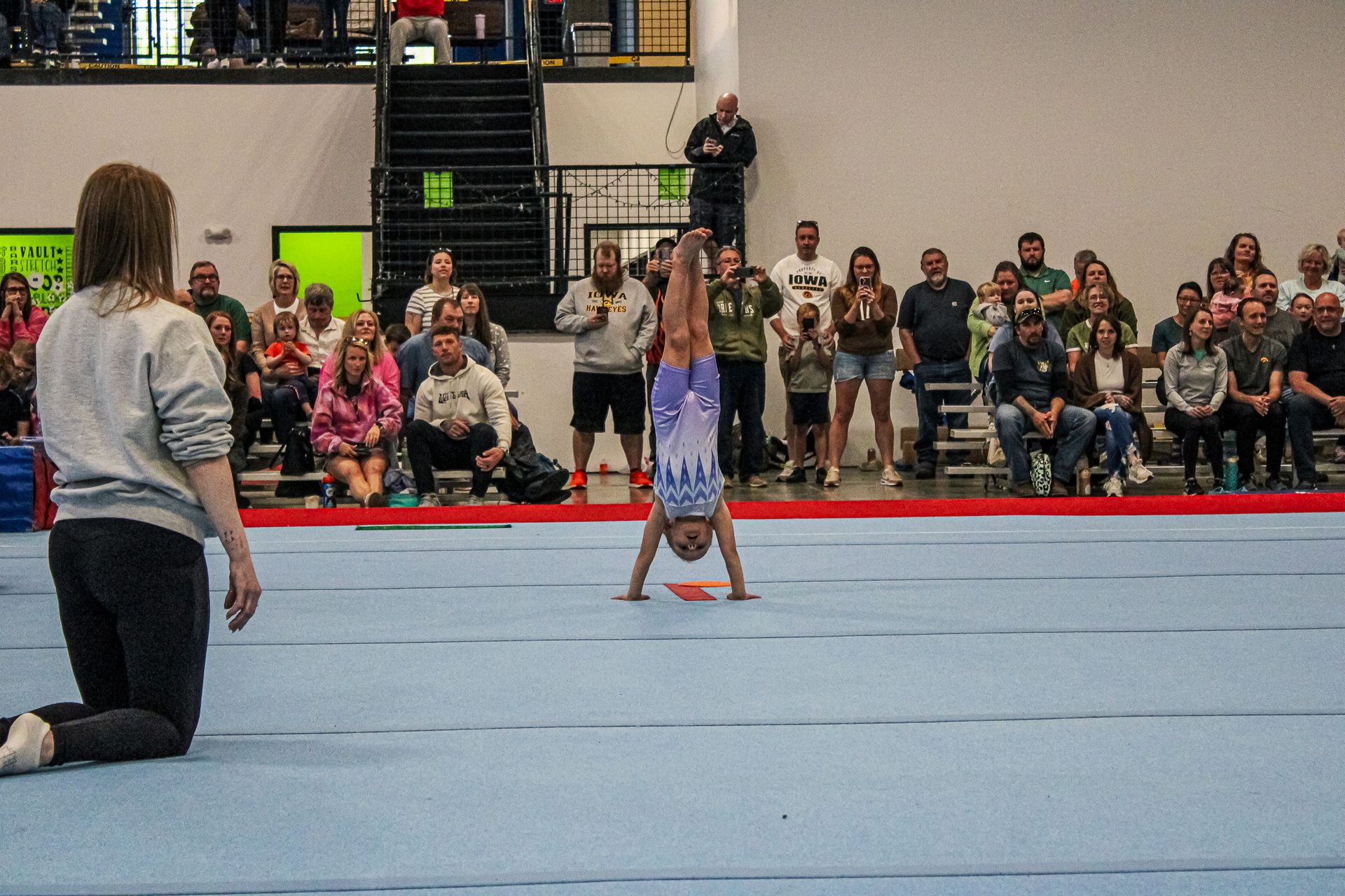 Gymnast performing a handstand on a blue mat while a coach kneels nearby at an indoor competition.