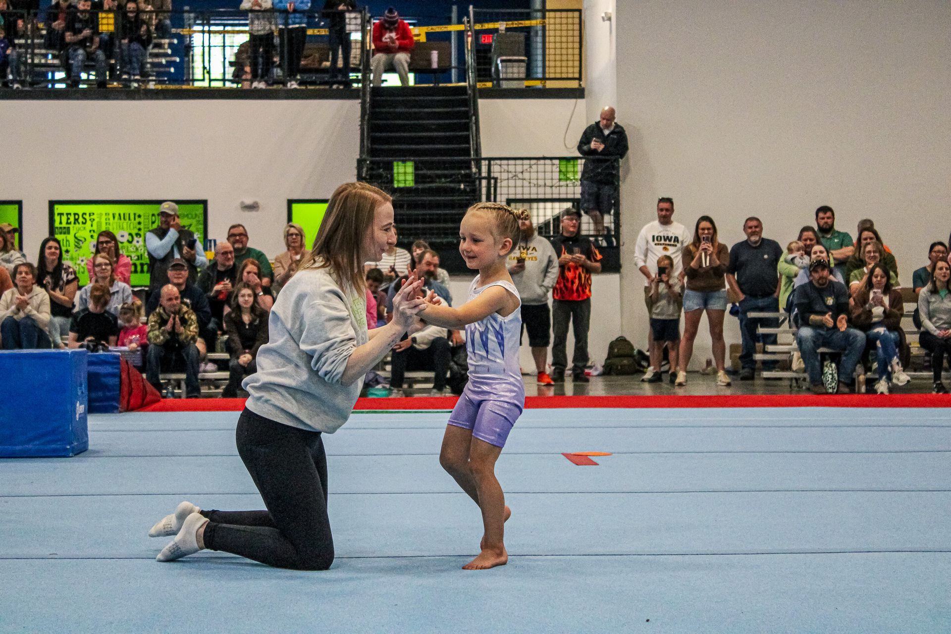 Coach kneels to support a young gymnast during a floor routine in an indoor gymnasium.