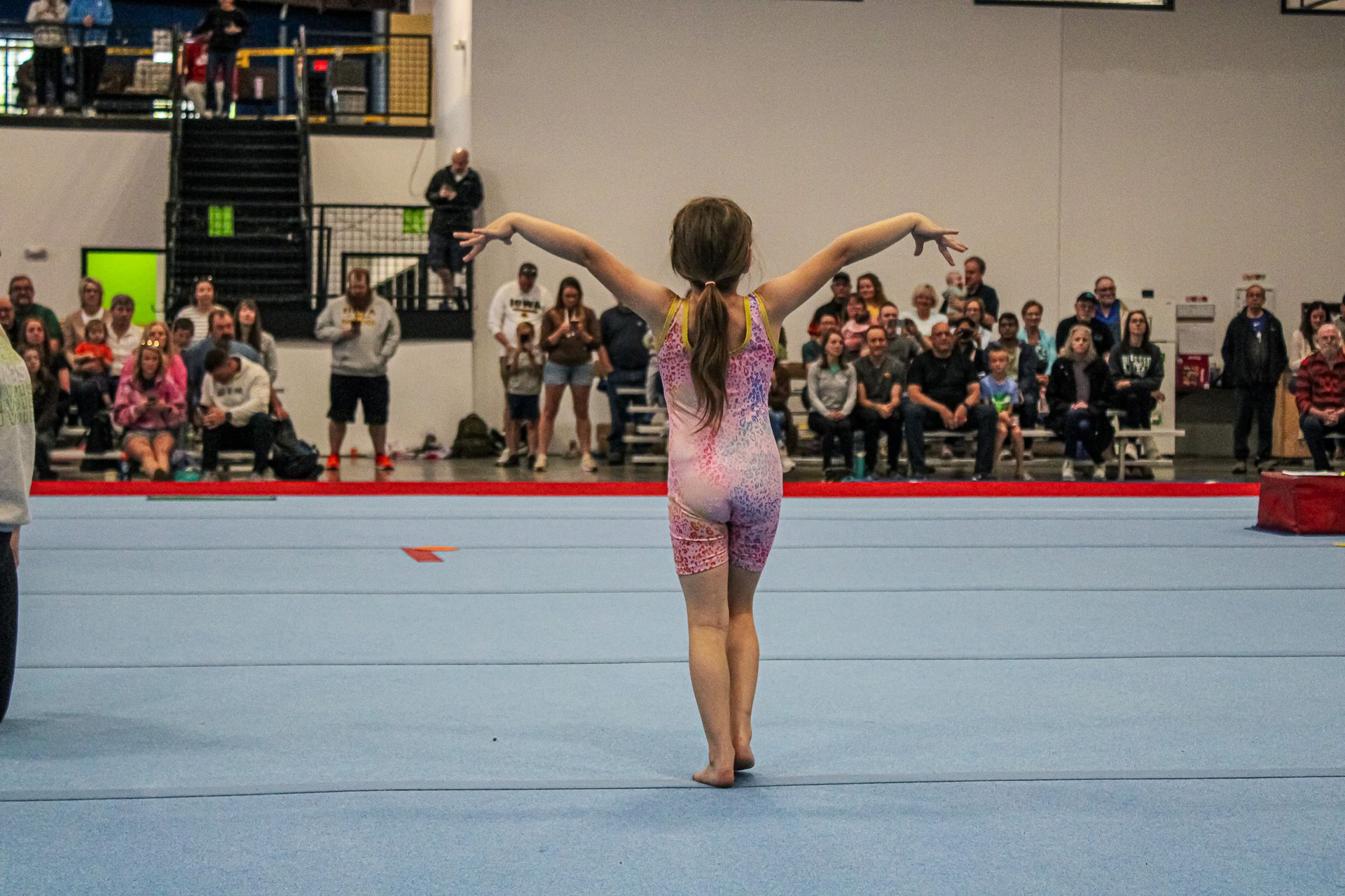 Gymnast in a pink leotard poses on a blue mat in an indoor competition arena.