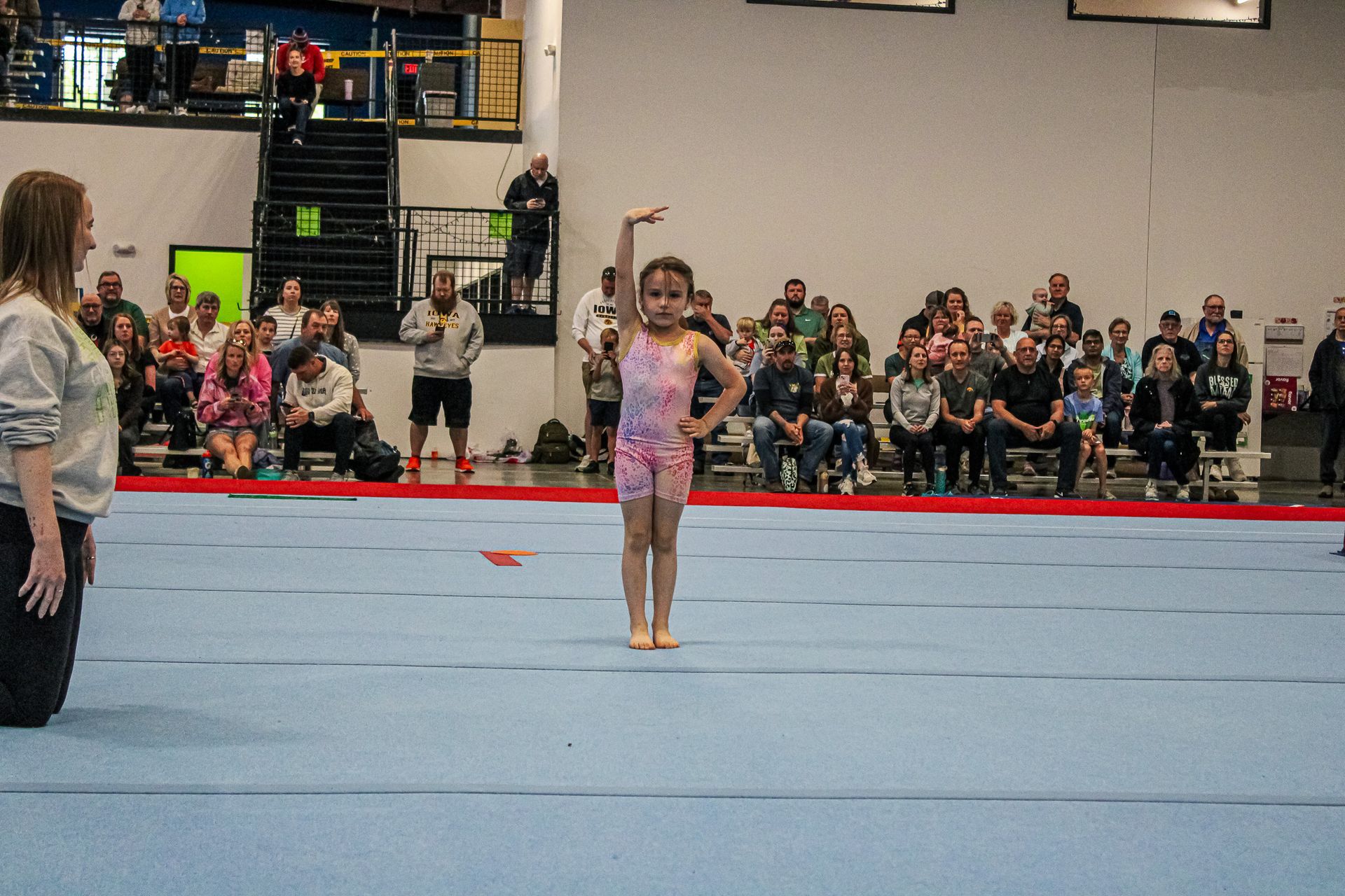 Gymnast standing on a blue mat in a gymnasium, with spectators seated in the background.