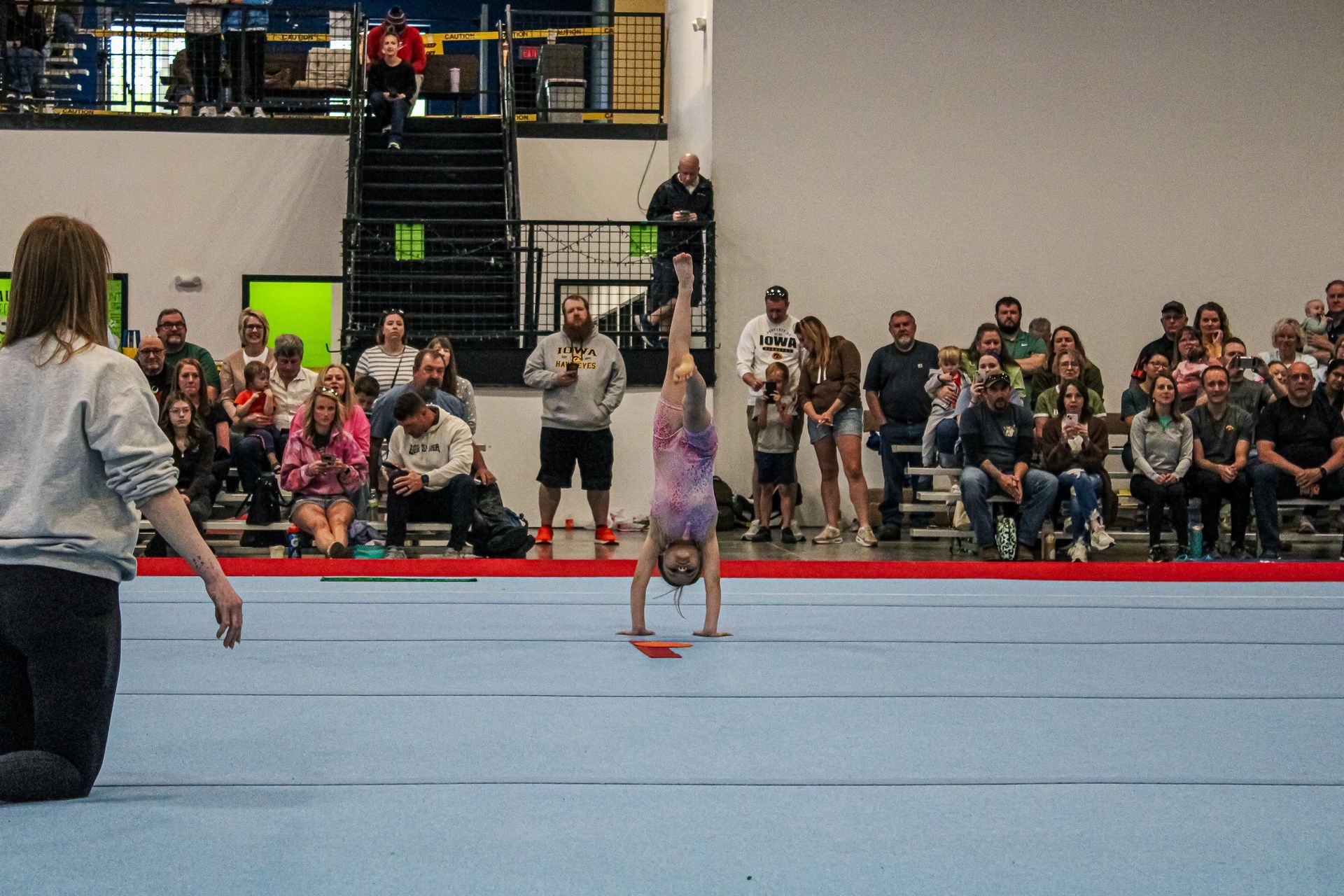 Gymnast doing a handstand on a blue mat in a gym, watched by an audience.