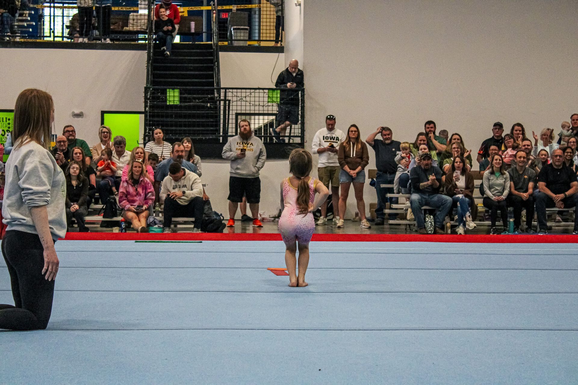 Child in pink leotard stands on a gym floor before an audience in a sports arena.