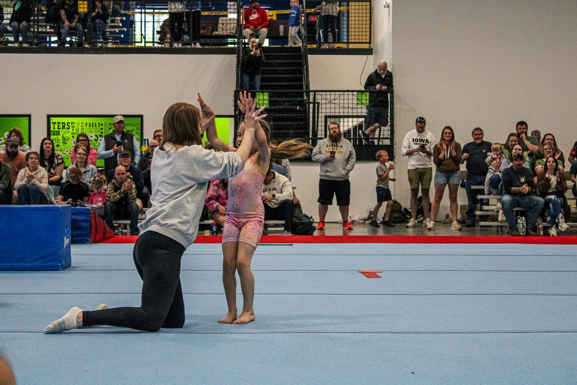 Coach kneels to assist a child gymnast on a blue mat during a gym performance, with spectators in the background