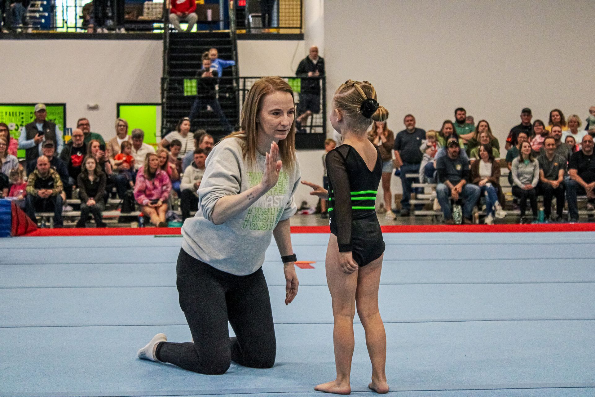 Coach kneels beside a gymnast on a blue mat in an indoor gymnasium with spectators in the background